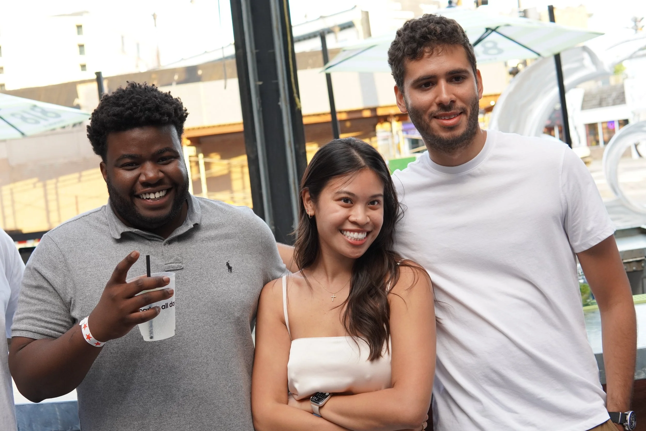 Three friends standing together outdoors, smiling for a photo. The man on the left has dark curly hair, a beard, is wearing a light gray polo shirt, and holding a drink. The woman in the middle has long dark hair, is wearing a white top, and has a sm