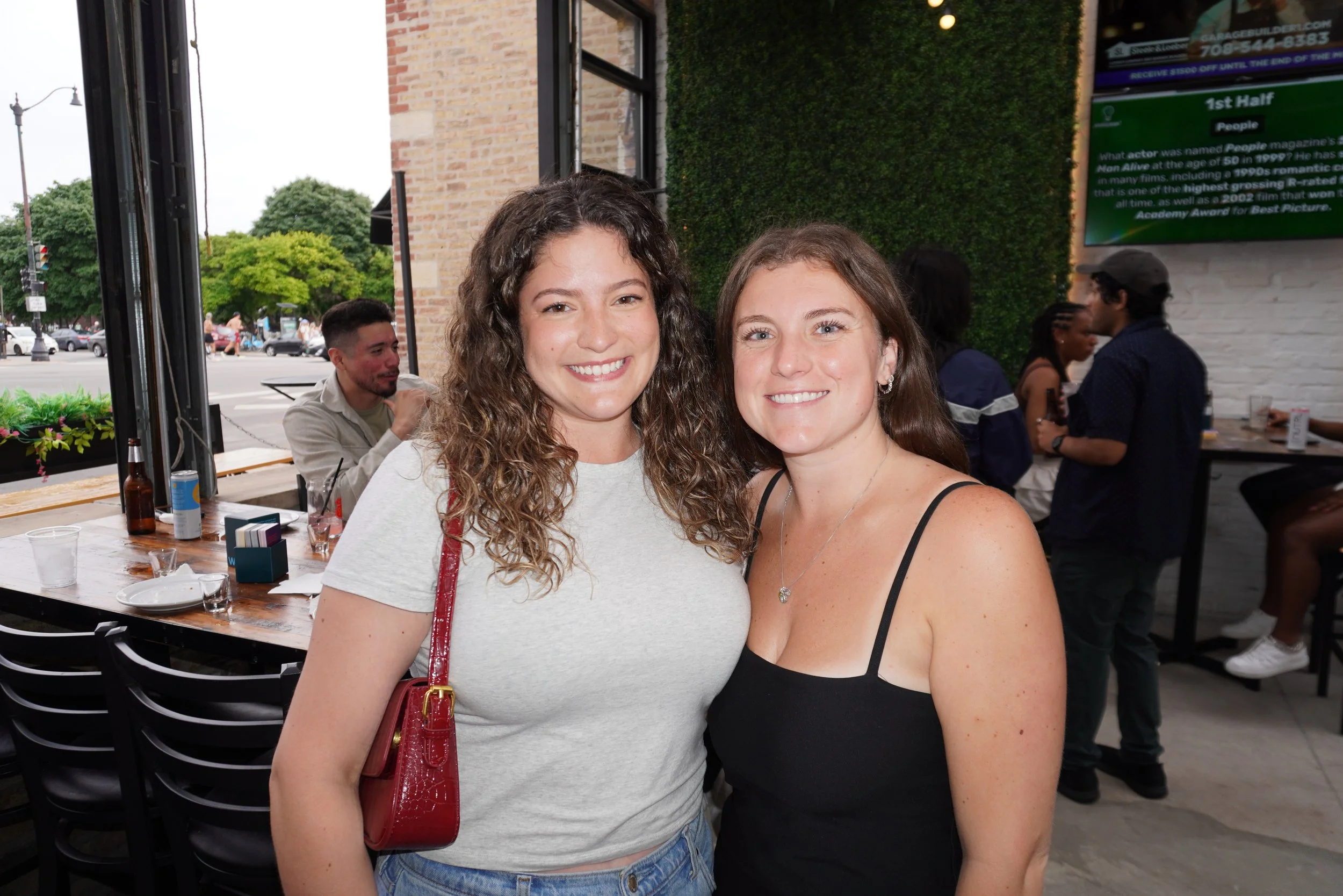 Two women smiling and posing together inside a restaurant or cafe, with other people in the background, and a view of the street outside.
