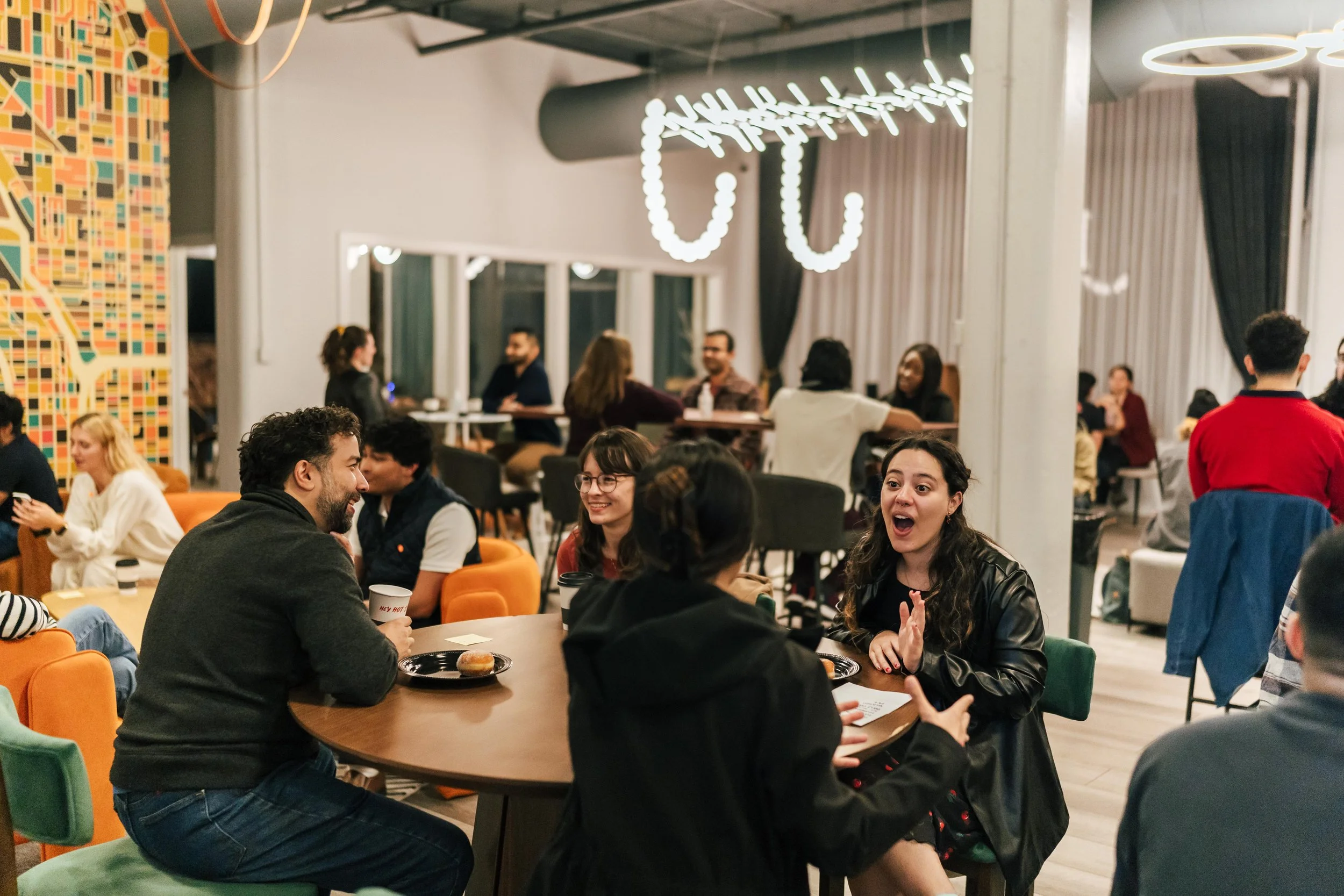 People socializing and chatting in a modern, lively indoor cafe or lounge, with colorful chairs, artwork, and contemporary lighting.