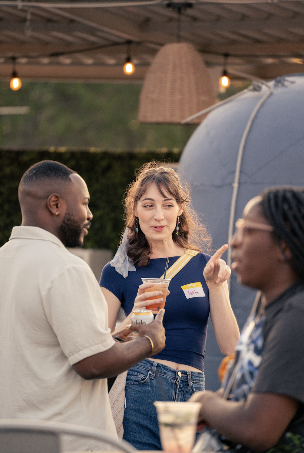 Three people talking at an outdoor gathering, one woman is gesturing while holding a drink and the two men listen, all are engaged in conversation.