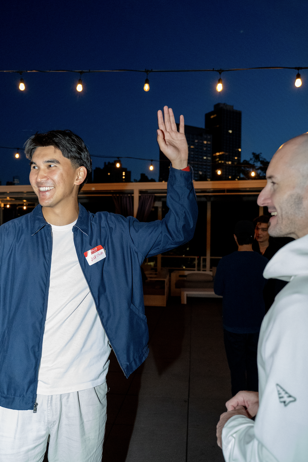 Two men smiling at each other during an outdoor evening event, one of them raising his hand for a high five, with string lights and city buildings in the background.