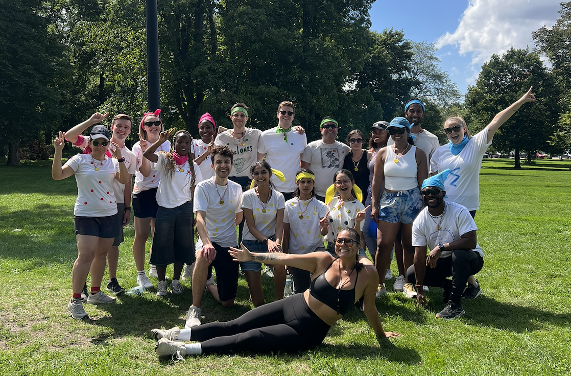 Group of people outdoors on a sunny day, some wearing white t-shirts with colored dots, posing for a photo in a park with trees and grass.