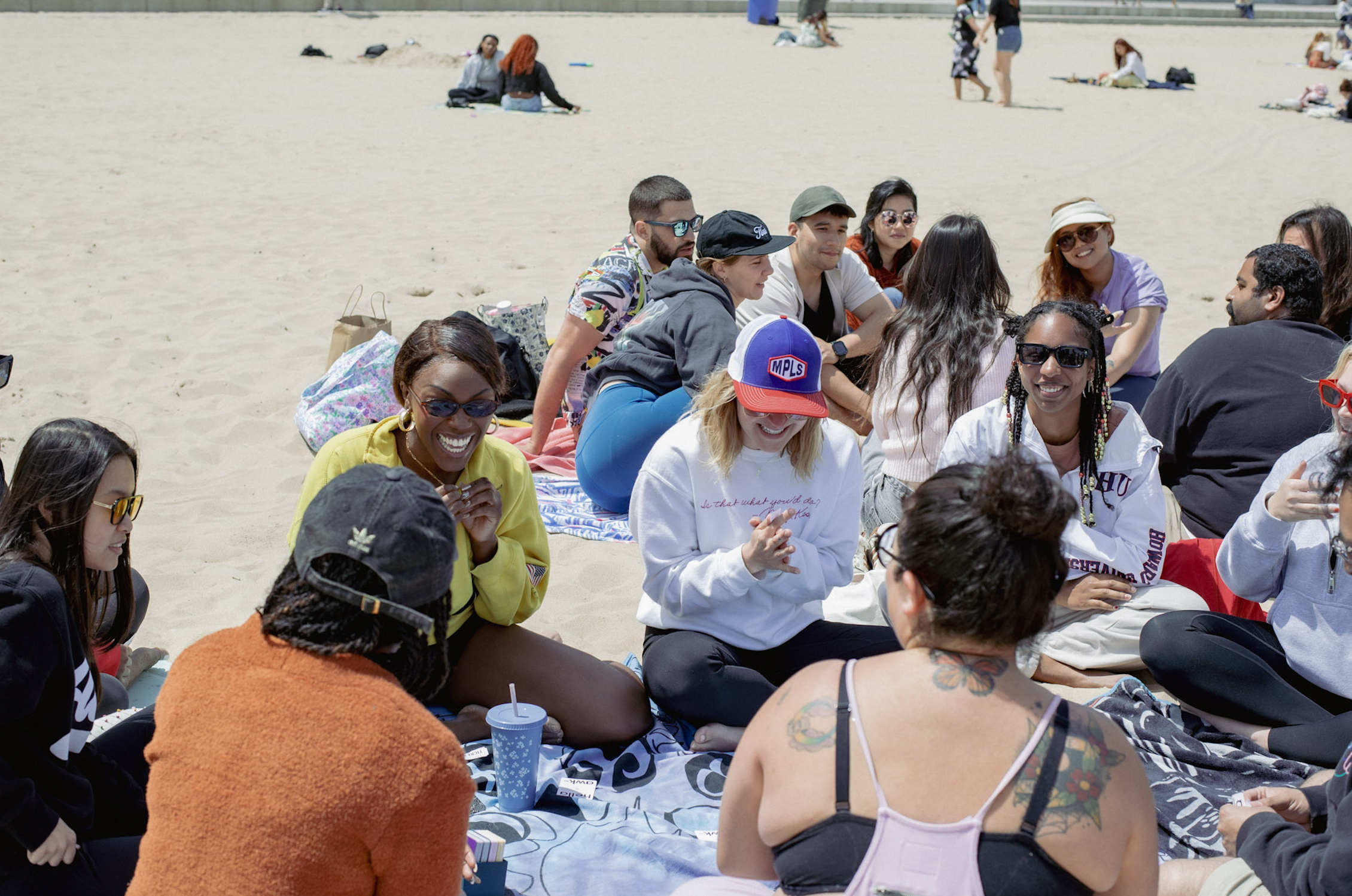 A group of people sitting on beach towels in a circle, smiling and chatting, with others in the background on the sandy beach.
