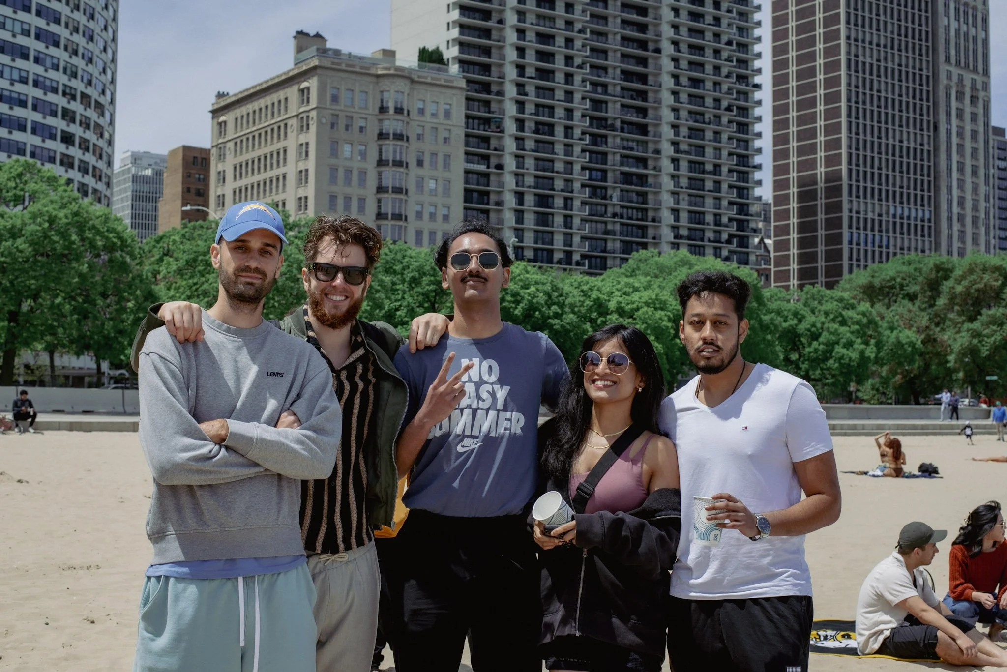 Group of five diverse friends standing together at a city park on a sunny day, smiling and posing for a photo, with high-rise buildings and green trees in the background.