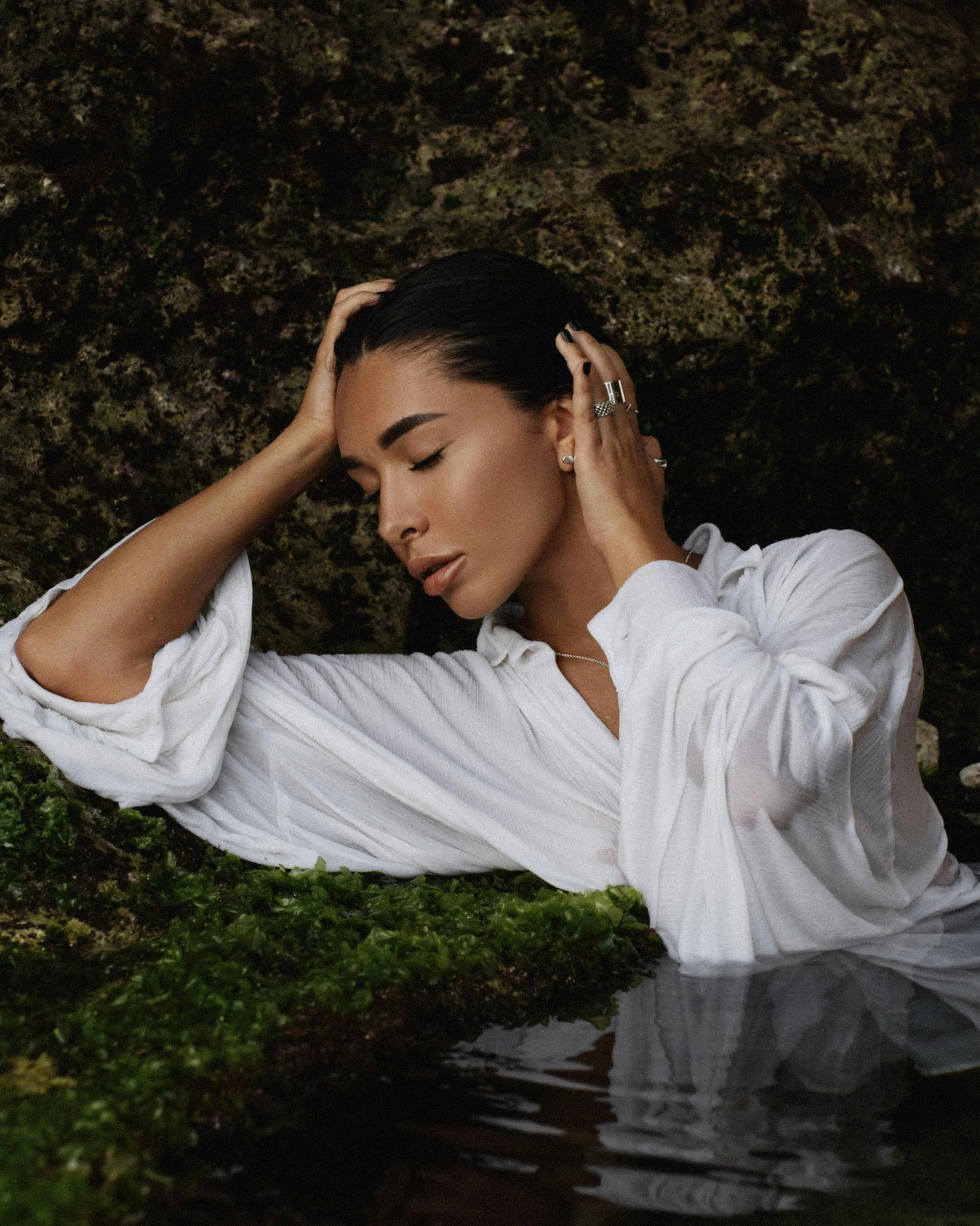 Une femme aux cheveux noirs, portant une chemise blanche, repose avec les yeux fermés contre une surface rocheuse recouverte de mousse, près de l'eau.