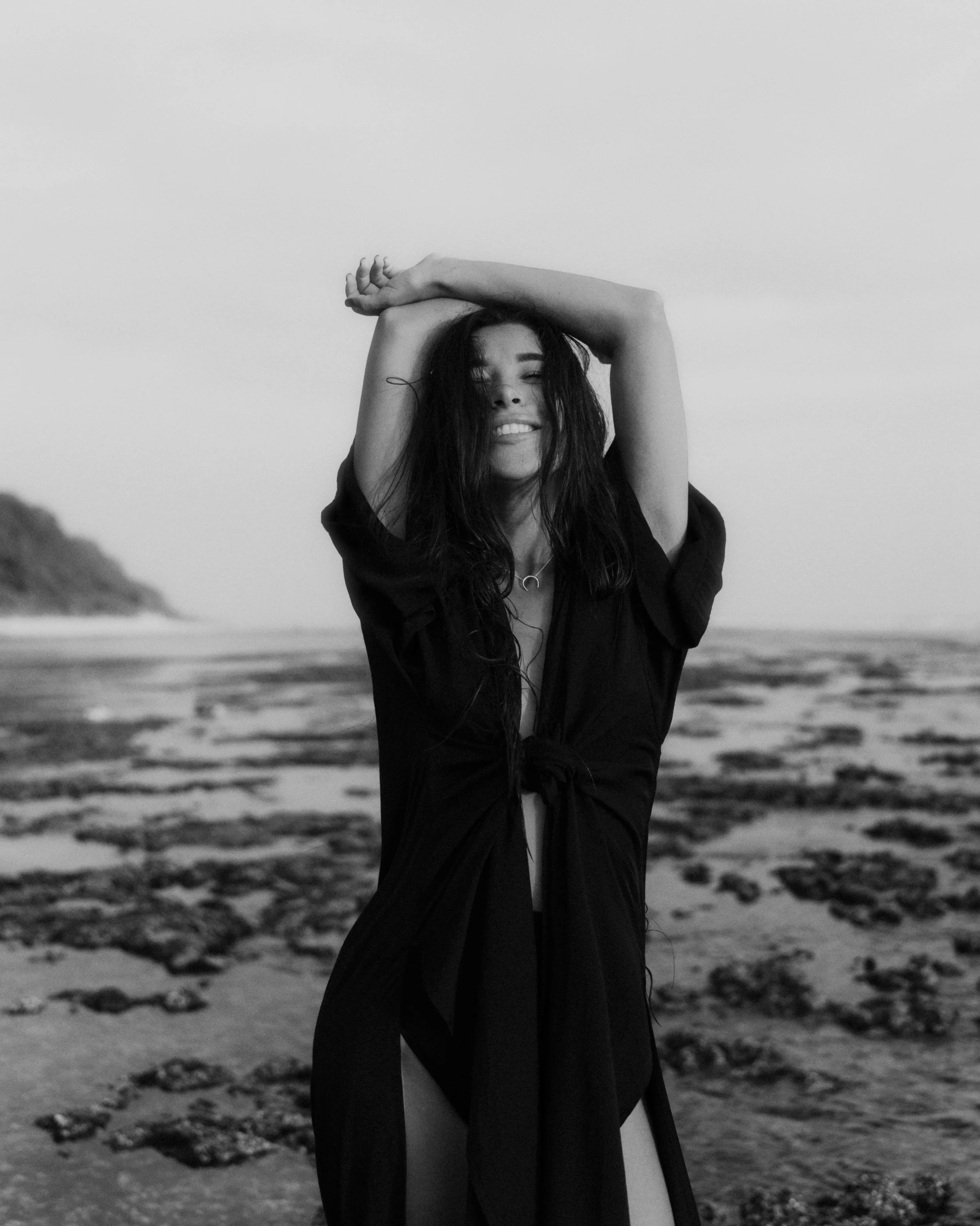 Une femme souriante avec les cheveux mouillés, portant une robe noire, sur la plage au coucher du soleil.