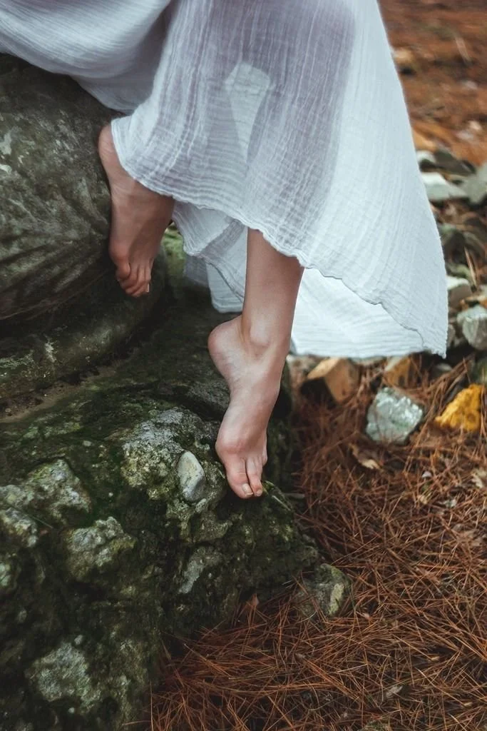 Une personne habillée d'une robe blanche est suspendue entre une roche et le sol, pieds nus, dans un environnement naturel avec des feuilles mortes.