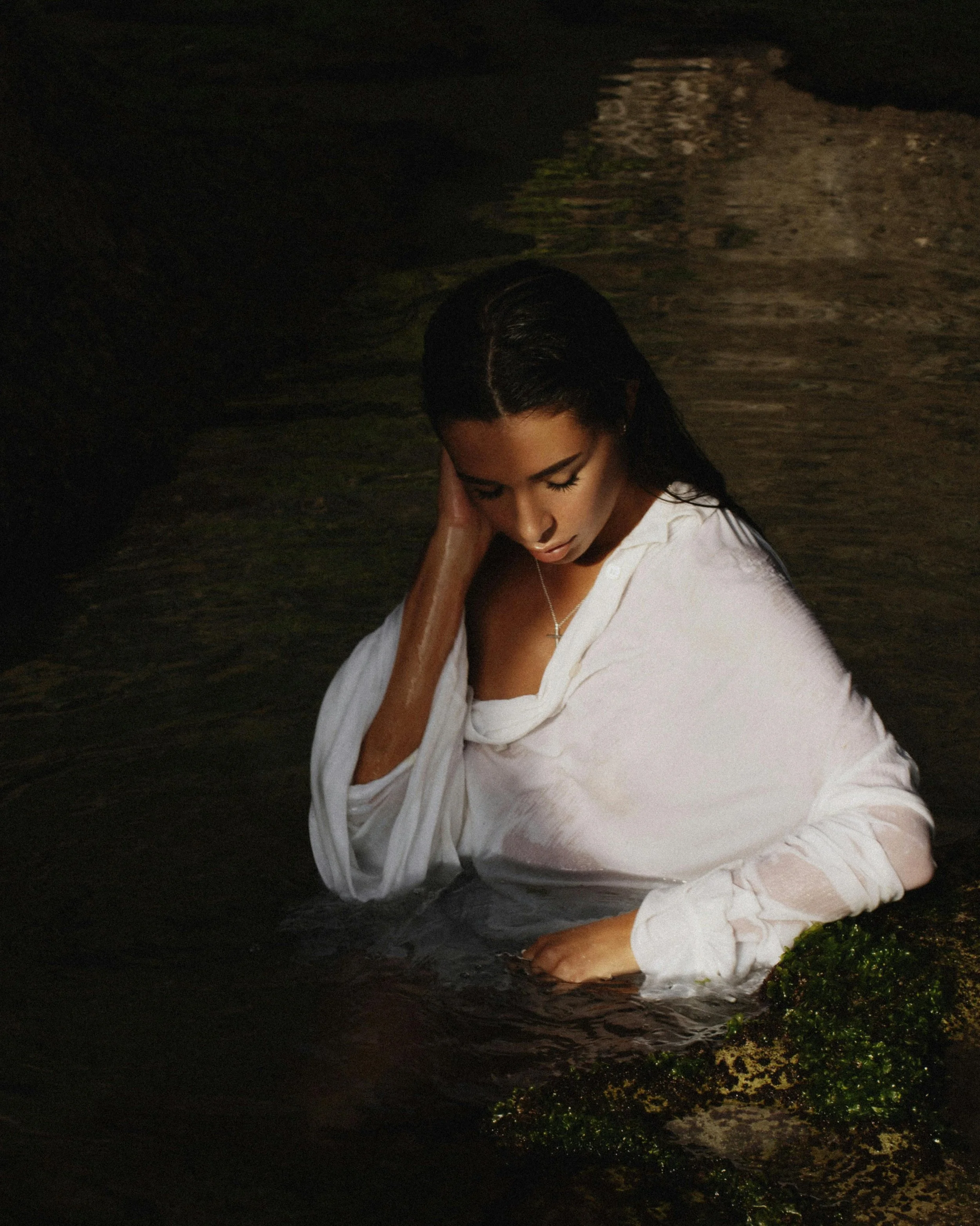 Une femme en robe blanche dans l'eau, avec des rochers et de la mousse verte, semble dans une grotte ou un endroit sombre.