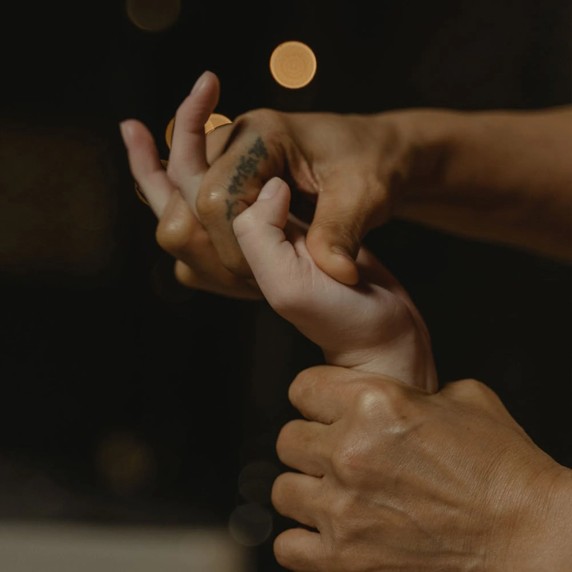 Deux mains qui se tiennent, une main est sombre et tatouée, l'autre est claire, dans un geste de poignée ou de présentation. Fond flou avec des lumières chaudes.