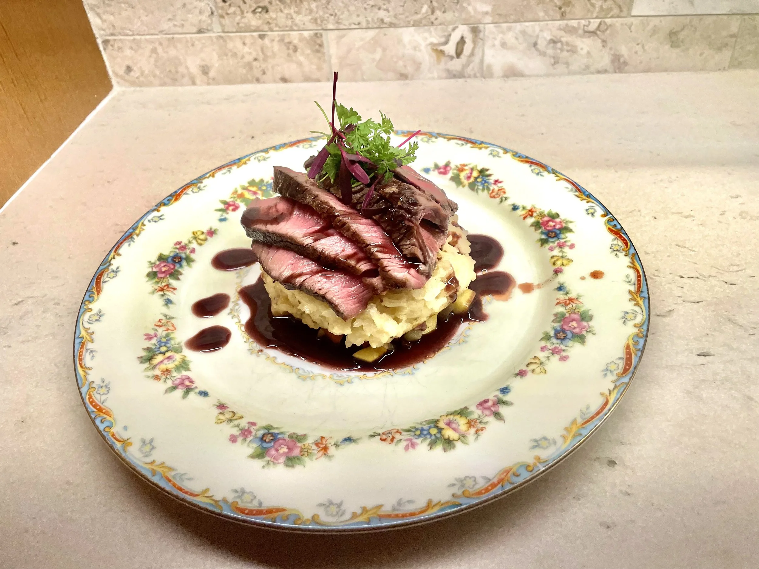 Slices of cooked steak on top of mashed potatoes with a garnish and a drizzle of sauce on a decorative floral plate.