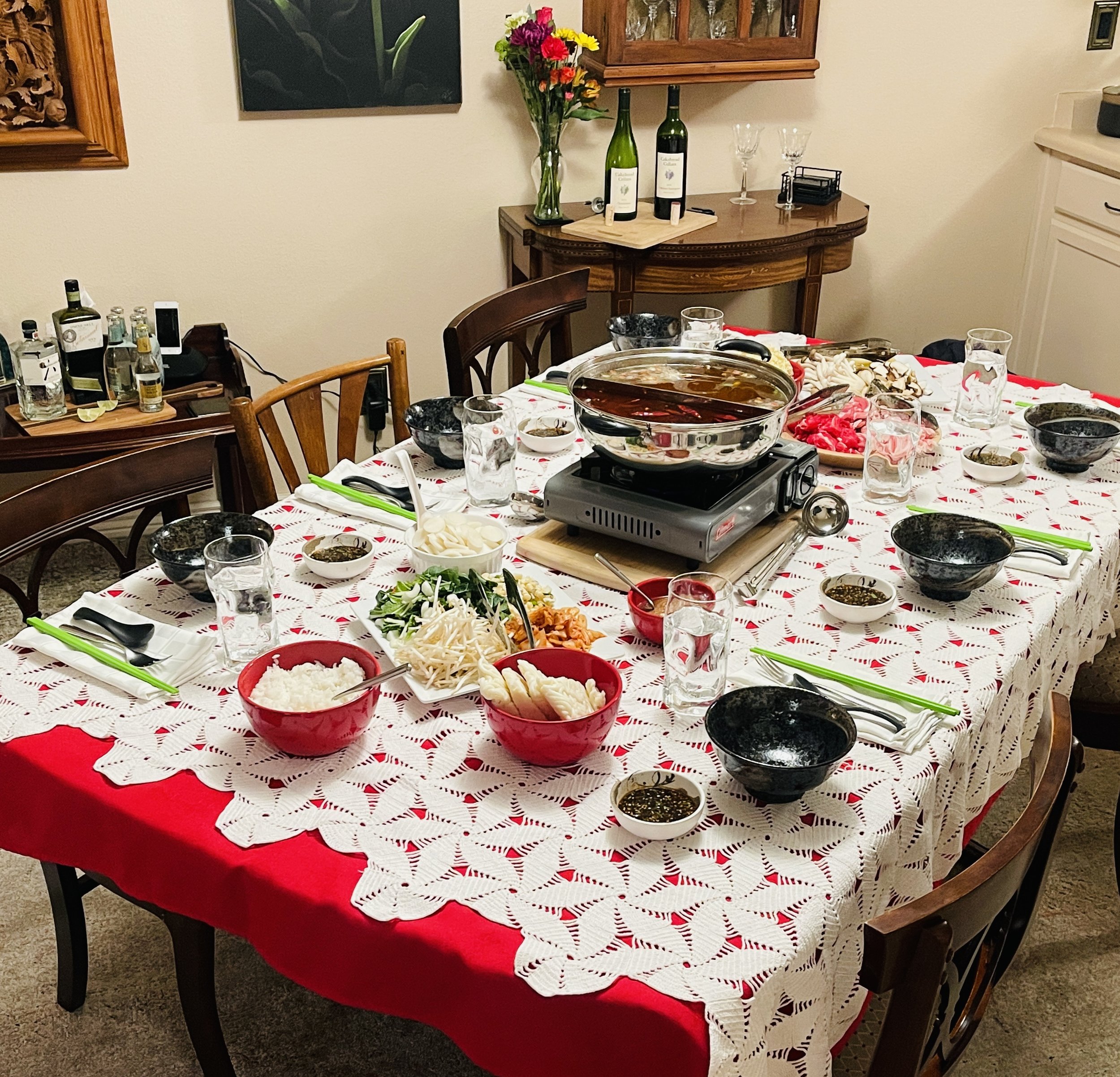 A dining table set for a hot pot meal with bowls of rice, vegetables, and sauces, surrounded by utensils and glasses, with a hot pot in the center on a portable stove, in a room with decorated walls and side tables.