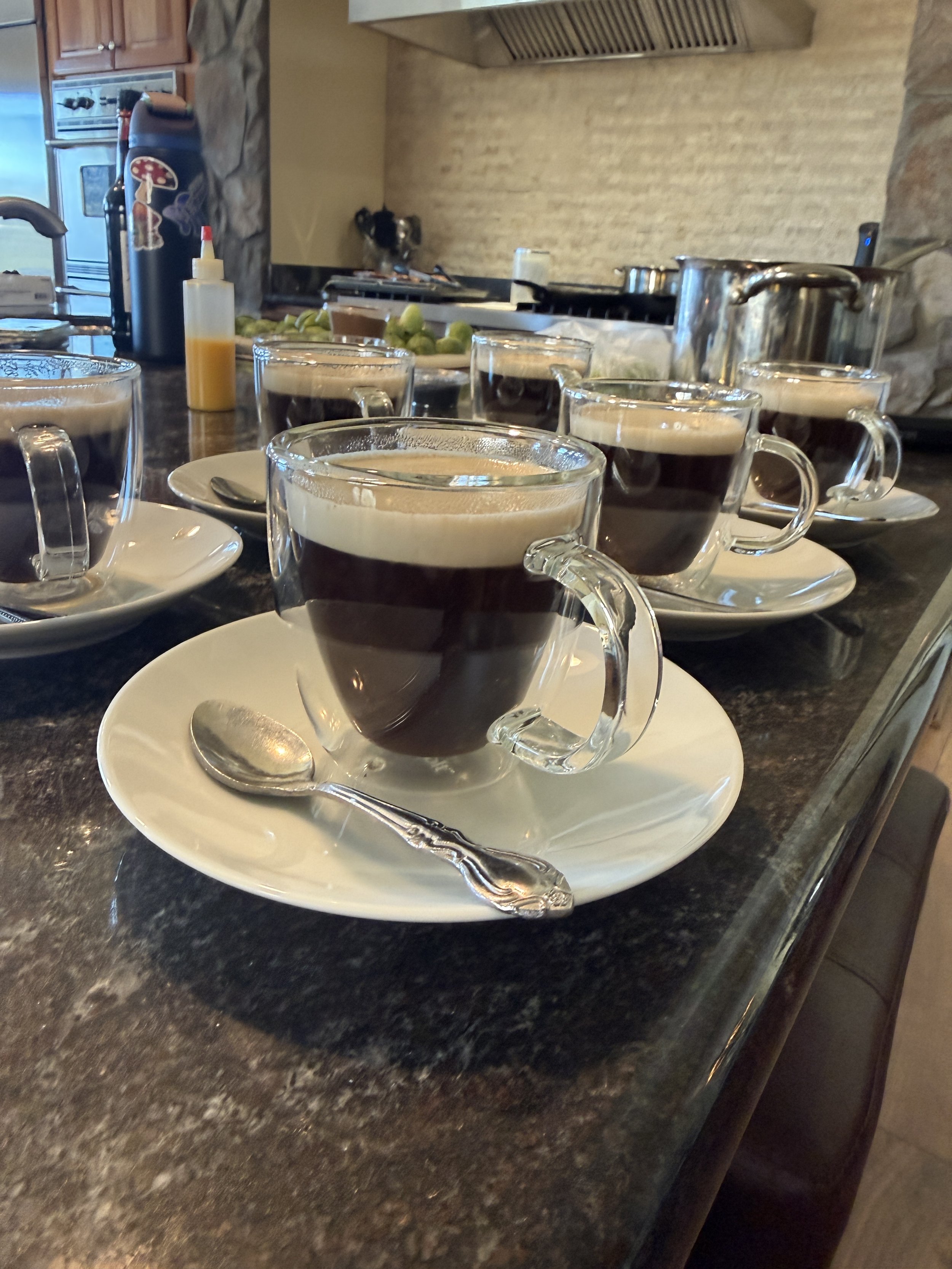 Six glass cups filled with black coffee placed on white saucers with small spoons on a dark granite kitchen counter.