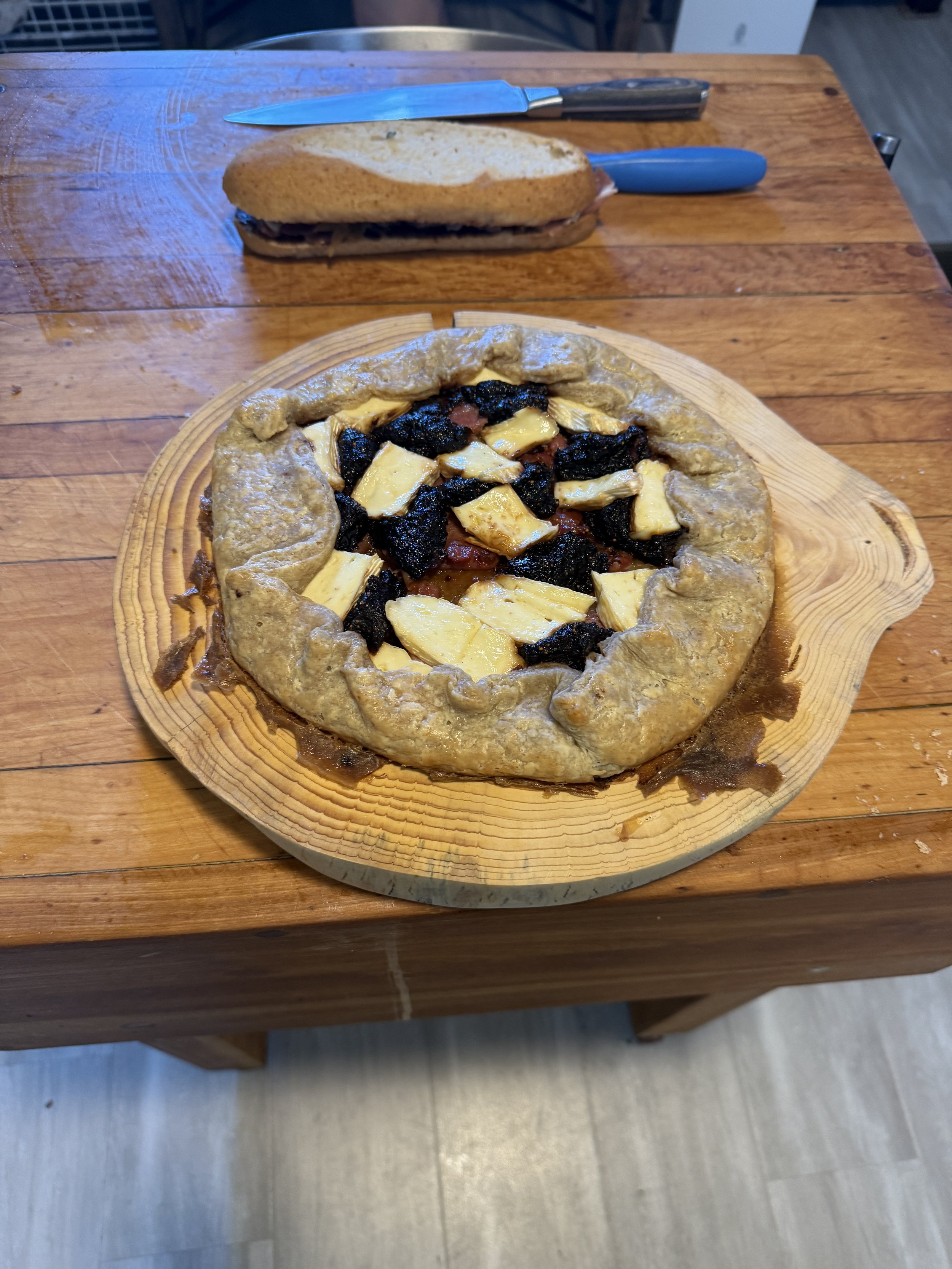 A pizza with a crust shaped like a pie, topped with cheese and blackened slices of what appears to be fruit or vegetables, sits on a wooden serving board. In the background, there is a sandwich with sliced bread, and a knife and a blue-handled utensi
