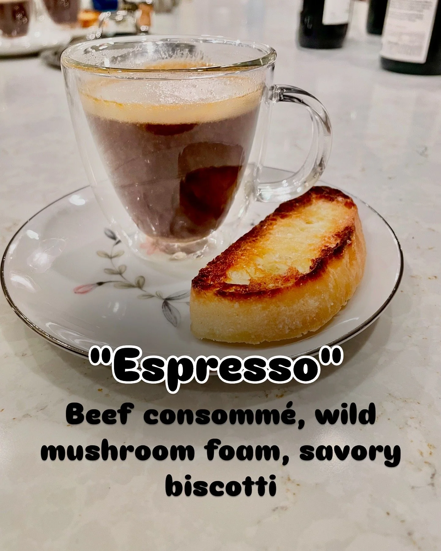 A clear glass cup of espresso with a layer of crema on top, placed on a white saucer with a floral design, next to a toasted bread slice, on a light-colored table.