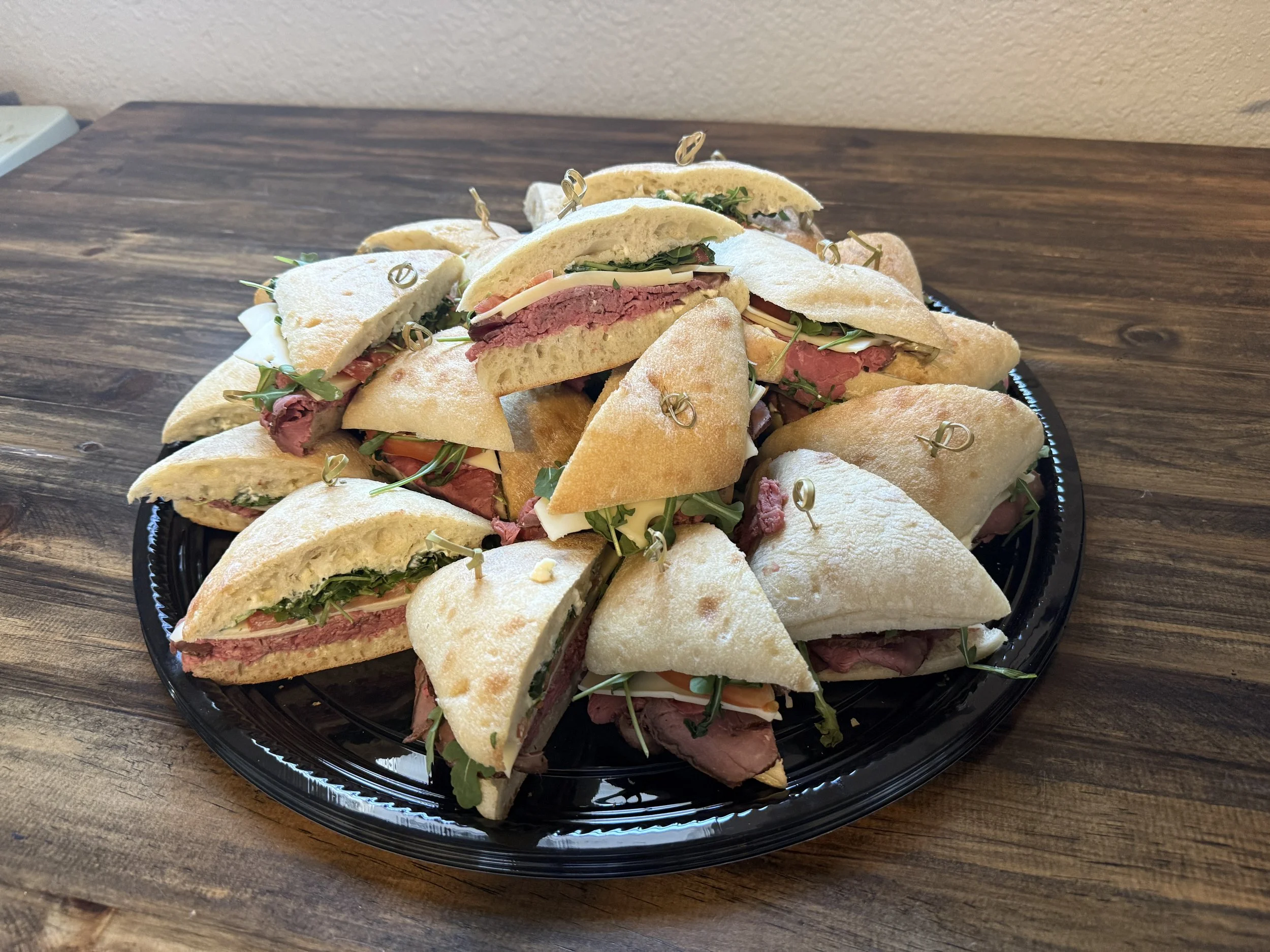 Tray of sliced sandwiches with roast beef, cheese, greens, and bread on a dark plastic platter placed on a wooden table.