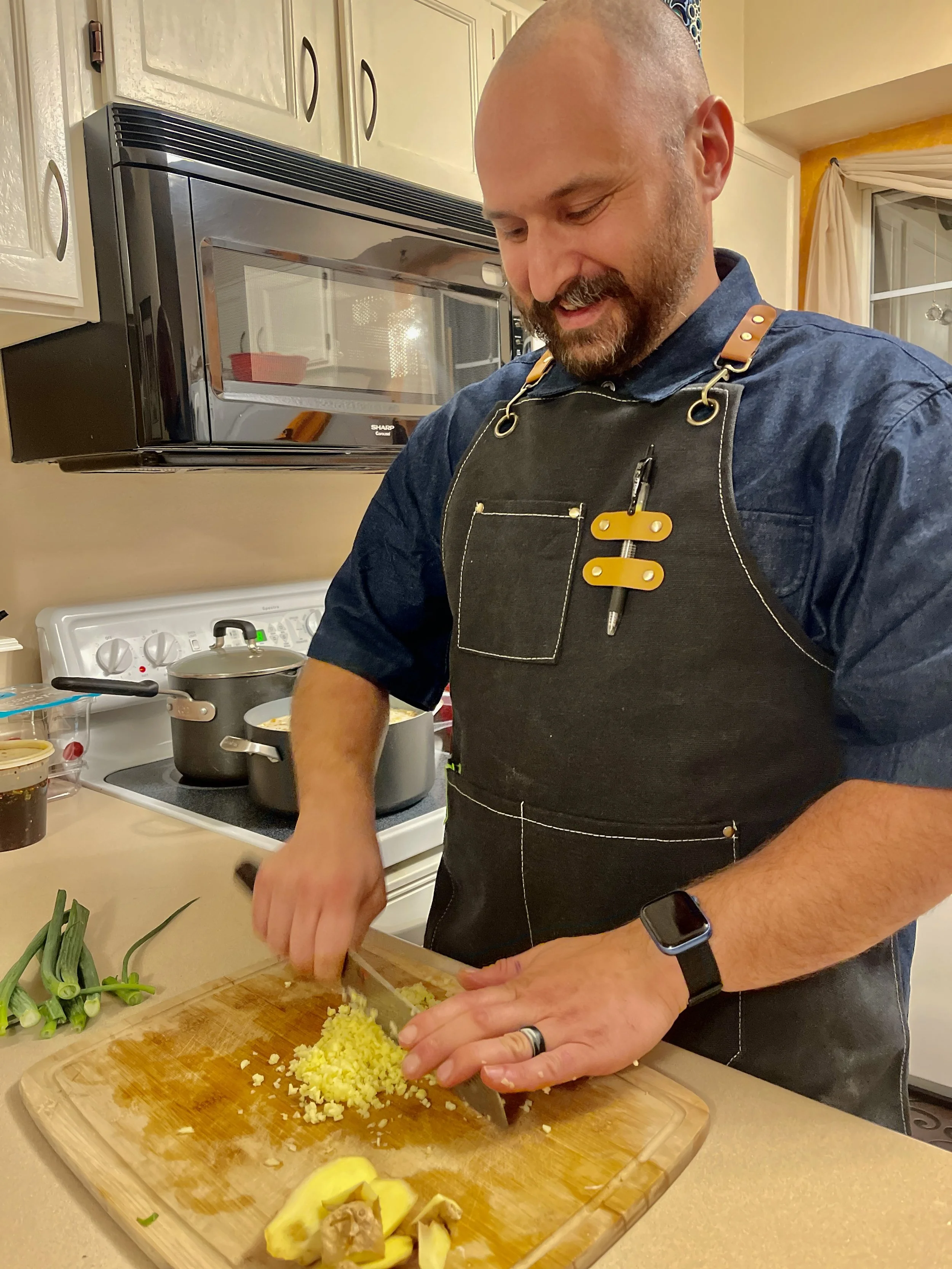 A man chopping yellow ginger on a wooden cutting board in a kitchen with a stove, microwave, and various cooking ingredients.