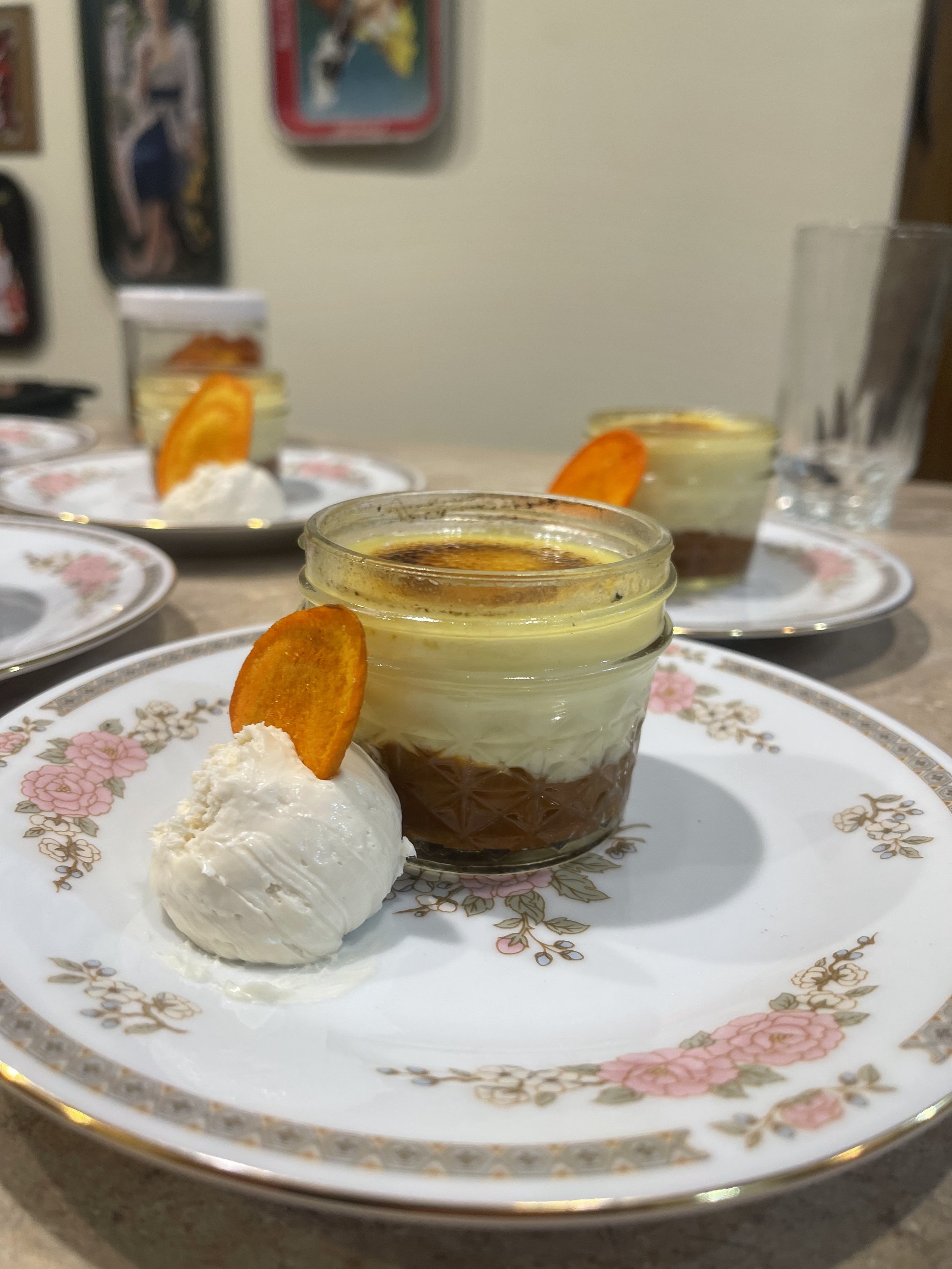 Close-up of a dessert served in a small glass jar, with layers of chocolate and cream, garnished with an orange slice and a dollop of whipped cream on a floral-patterned plate.