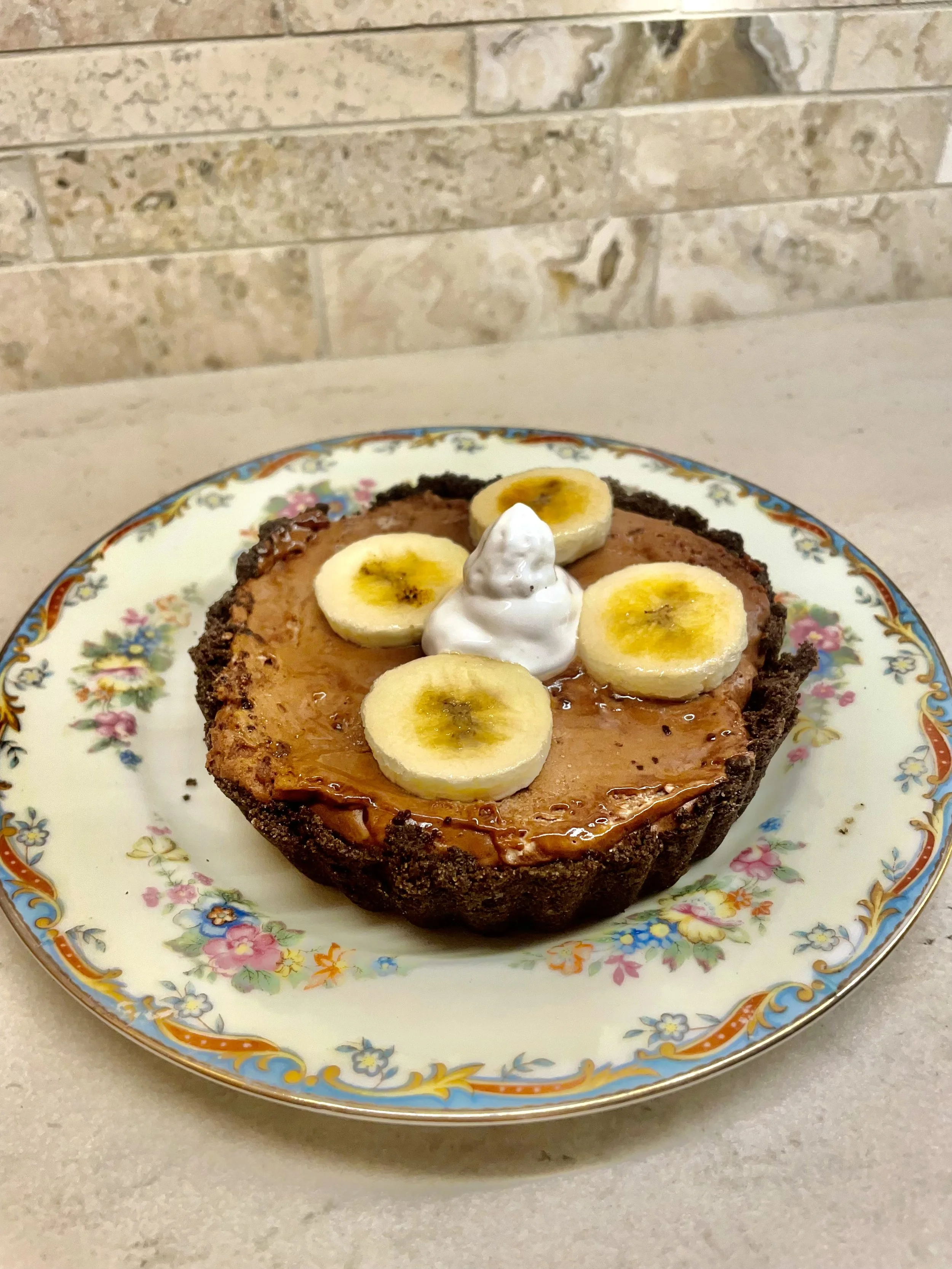 A banana cream pie topped with banana slices and a dollop of whipped cream on a decorative floral plate.