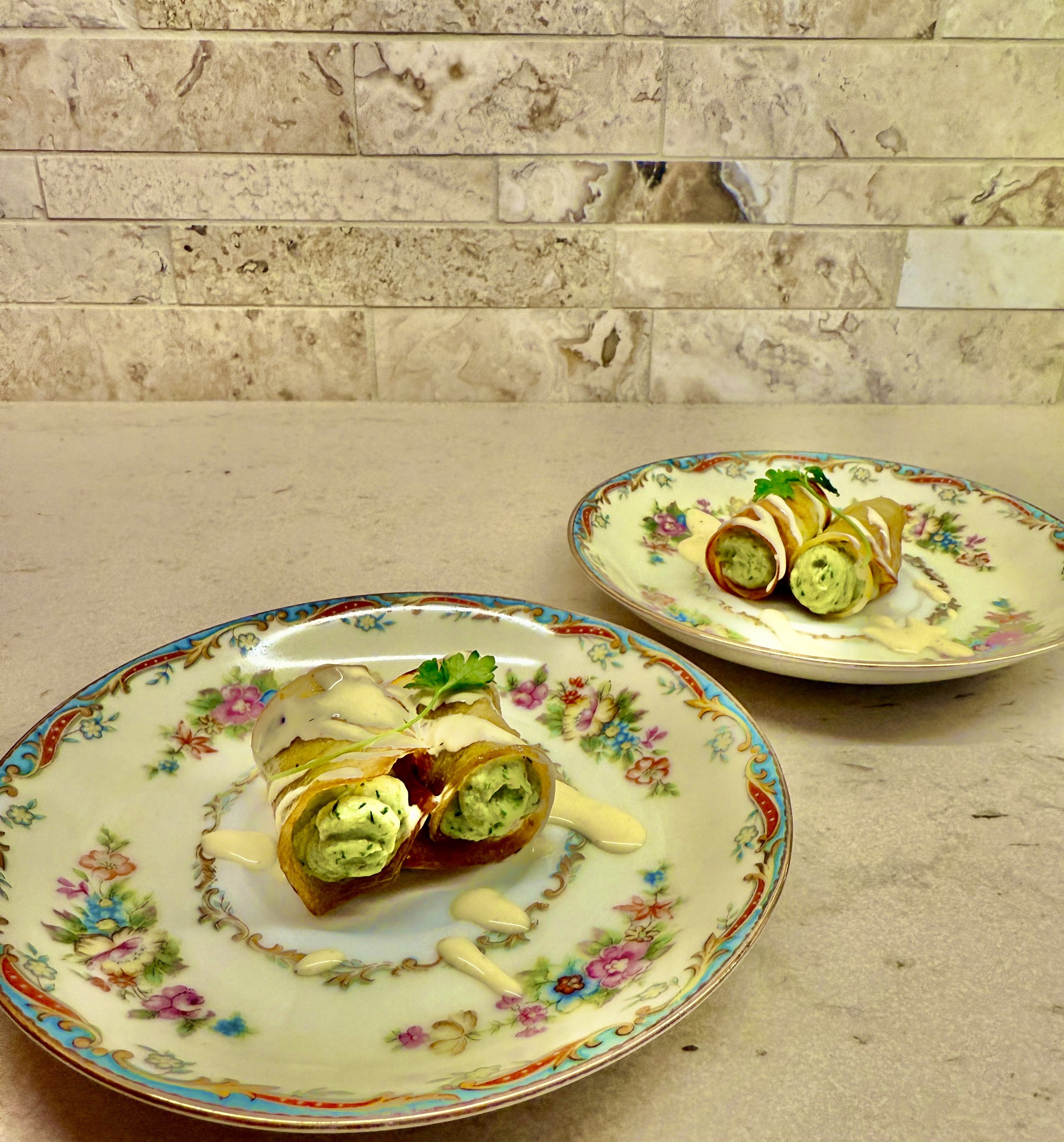 Two decorative plates with spring rolls drizzled with white sauce, garnished with cilantro, on a light-colored countertop with a brick wall background.