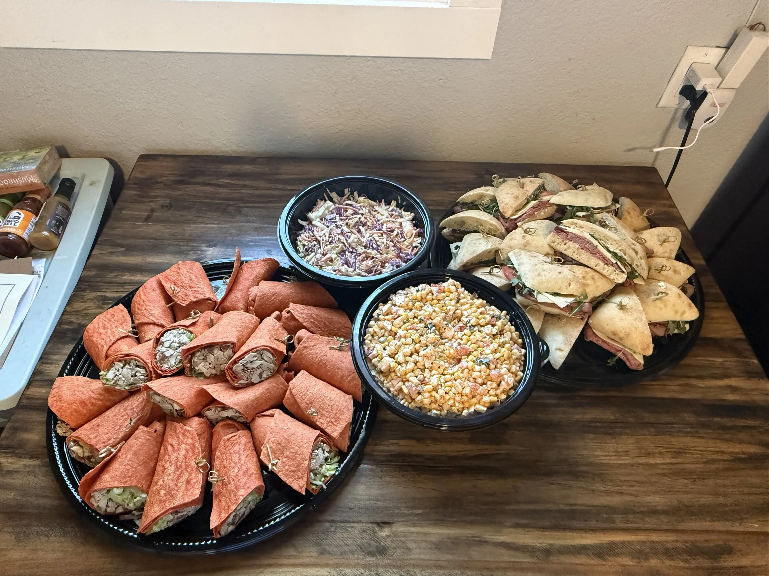 A spread of wrapped spring rolls, pasta salad, corn salad, and small sandwiches on a wooden table. There are bottles of sauce and condiments on a tray to the side.