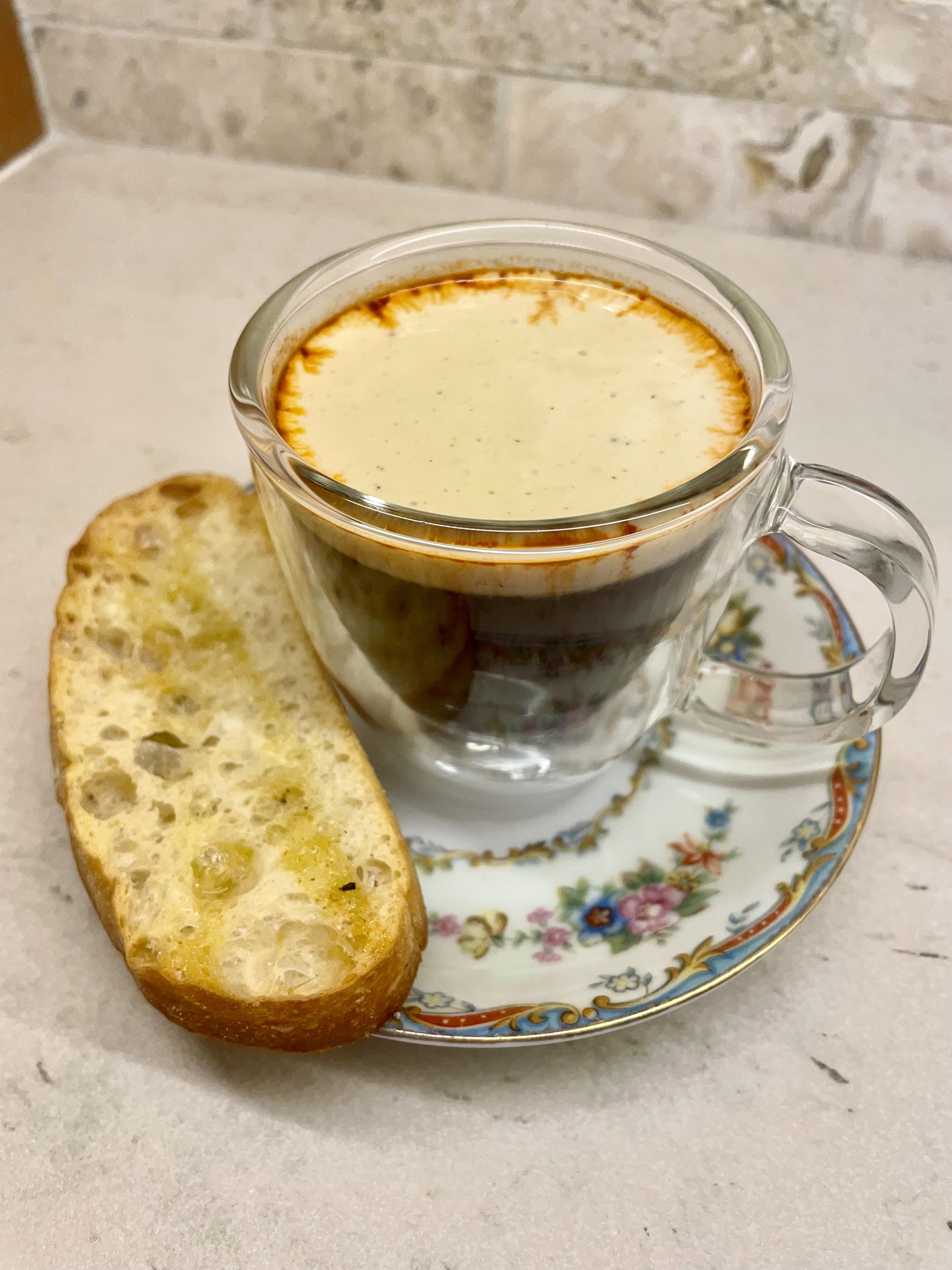 A clear glass cup of layered espresso topped with foam, served on a decorative floral plate with a slice of toasted baguette.
