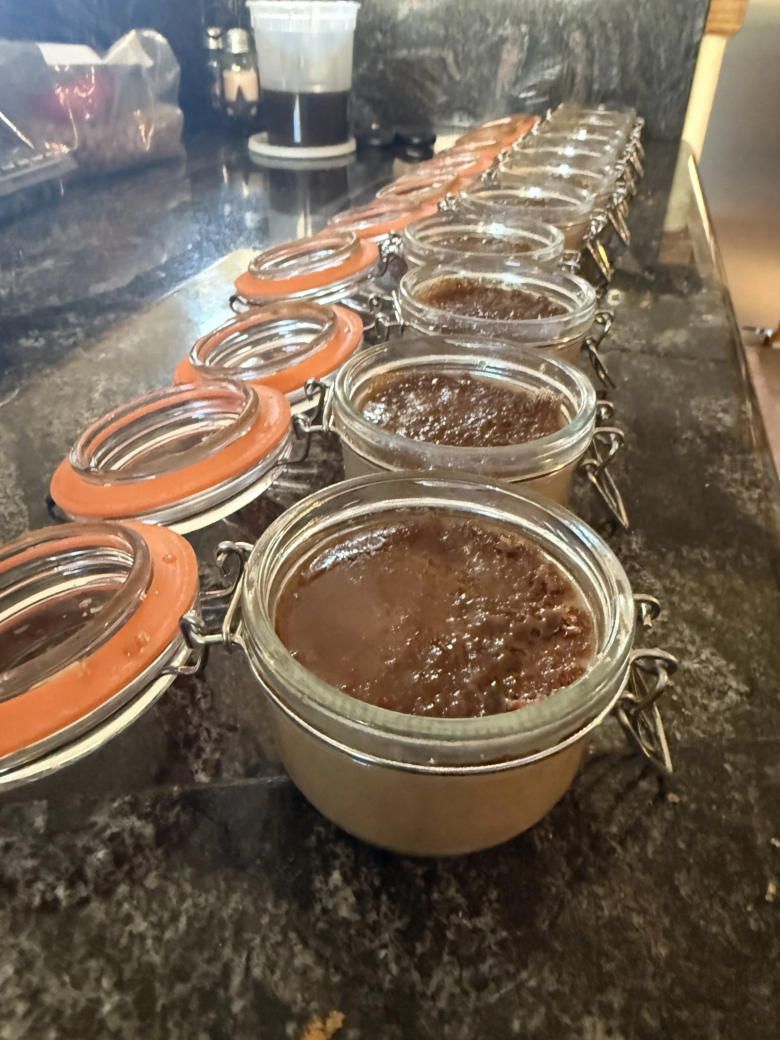 Multiple jars of homemade jam or preserve lined up on a dark countertop, with a glass of dark beverage and a salt shaker in the background.