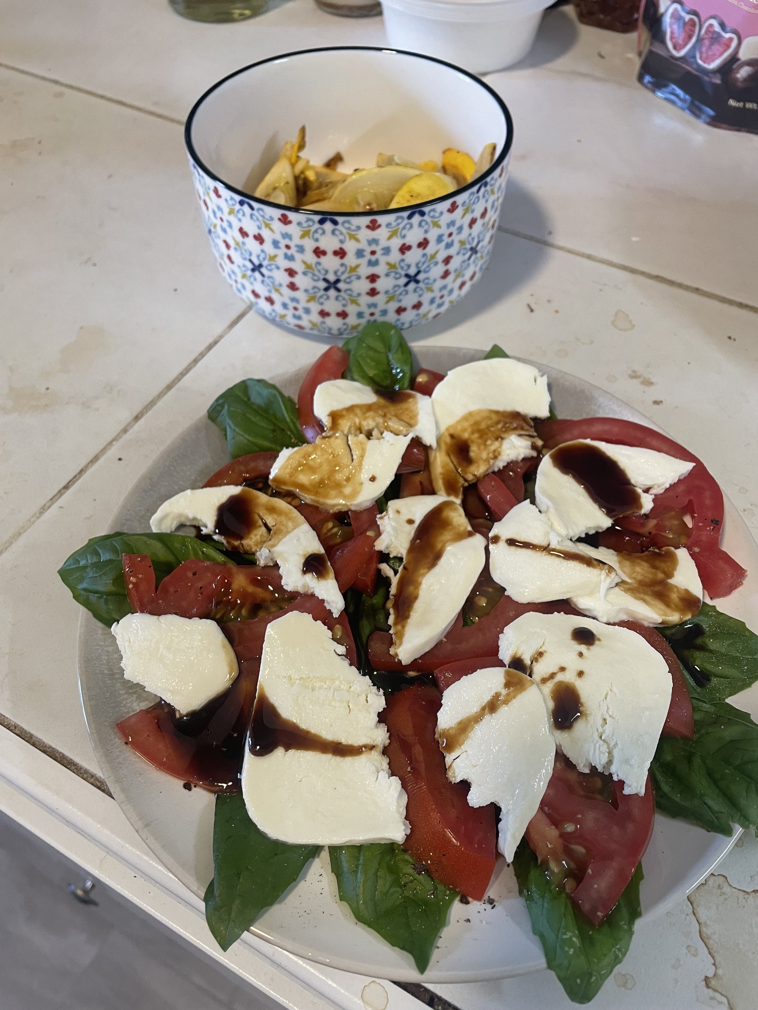 Caprese salad with sliced tomatoes, mozzarella cheese, basil leaves, balsamic glaze, and olive oil on a plate, with a bowl of artichoke hearts in the background.