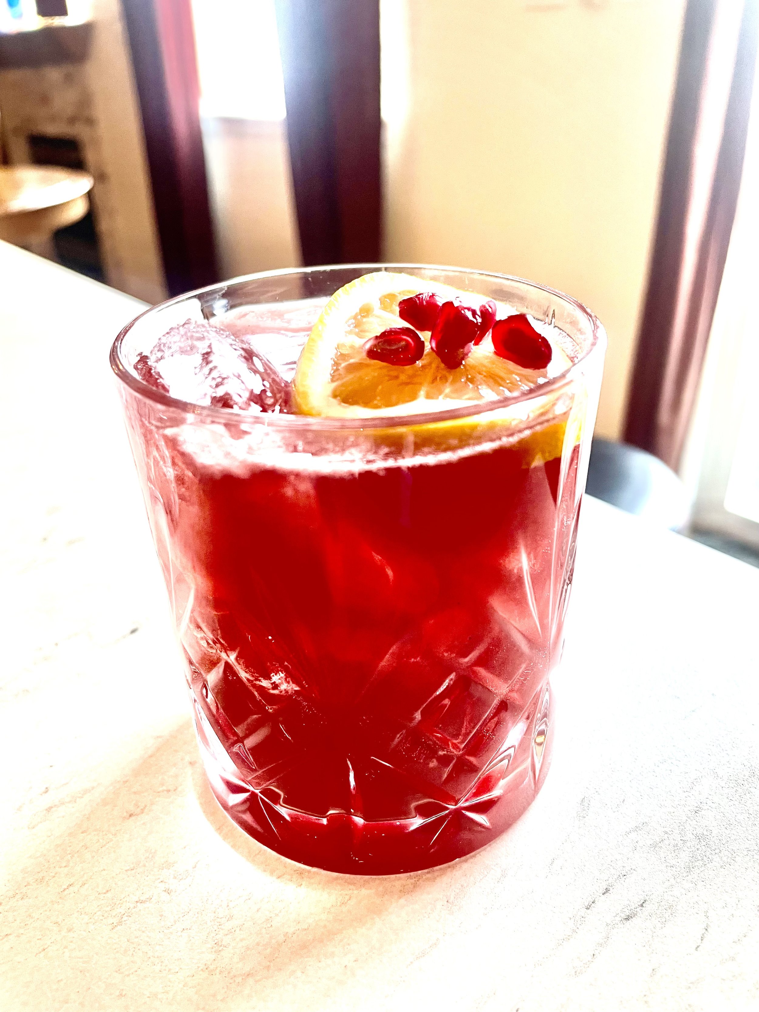 A glass of red beverage garnished with a lemon slice and pomegranate seeds, on a table near a window.
