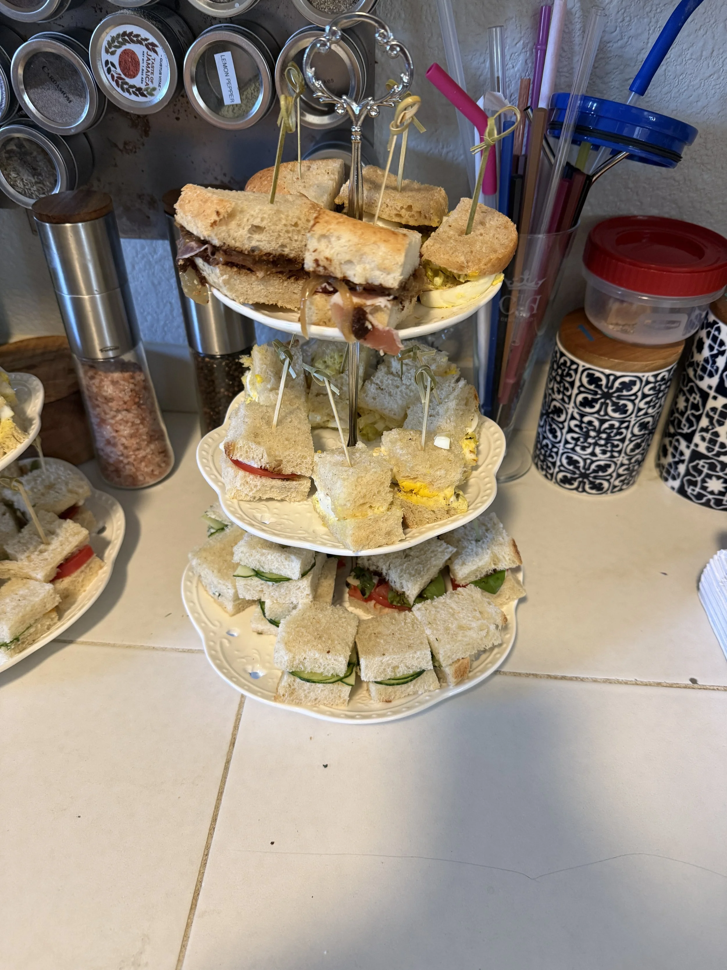 Three-tiered tray with assorted finger sandwiches and mini sandwiches, surrounded by various kitchen items and containers on a countertop.