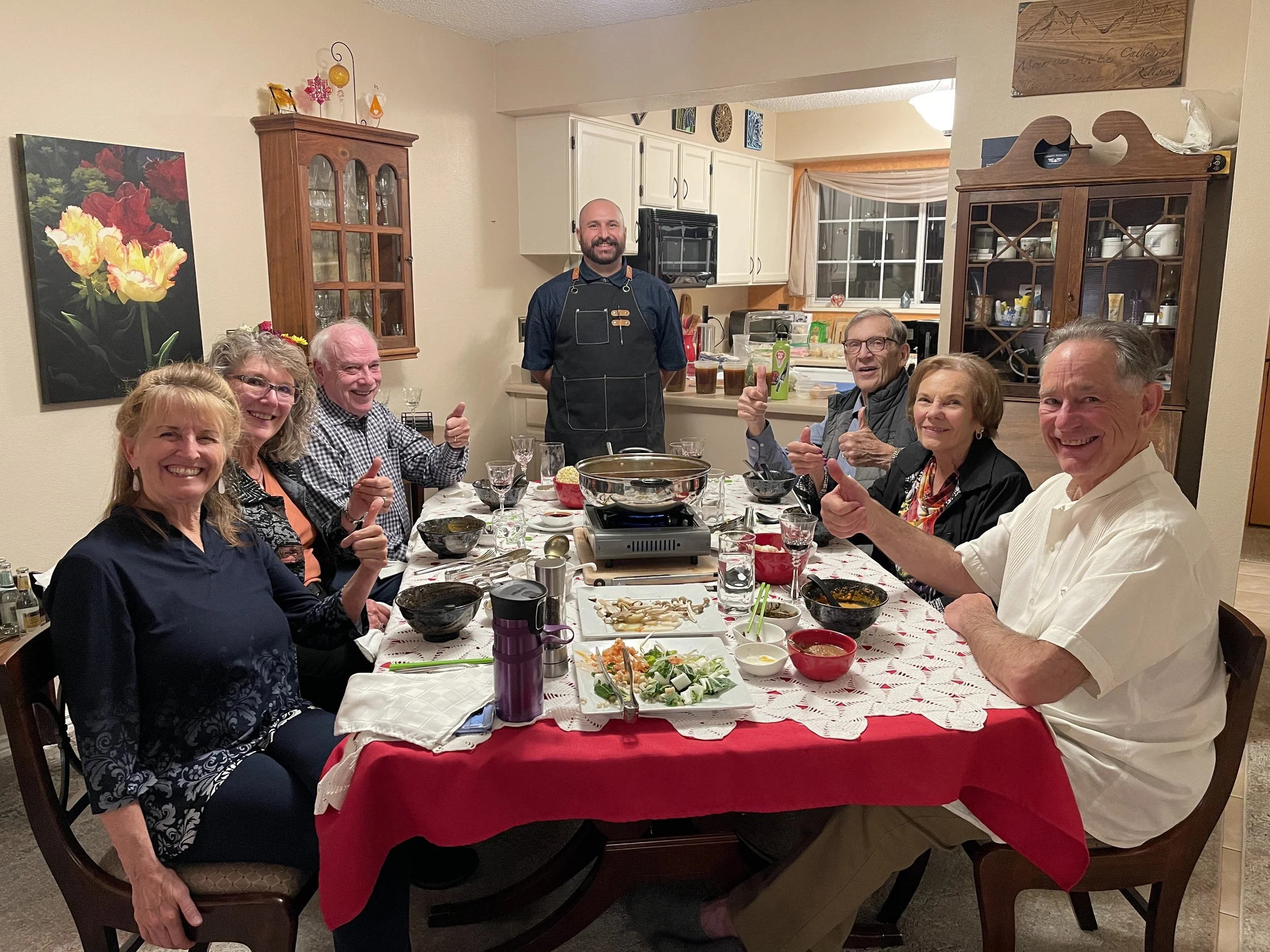 A group of seven people, six adults and one young adult, sitting around a dining table enjoying a meal, with a chef standing behind them in a kitchen, all smiling and giving thumbs up. The table is set with various dishes and drinks, and the room is 
