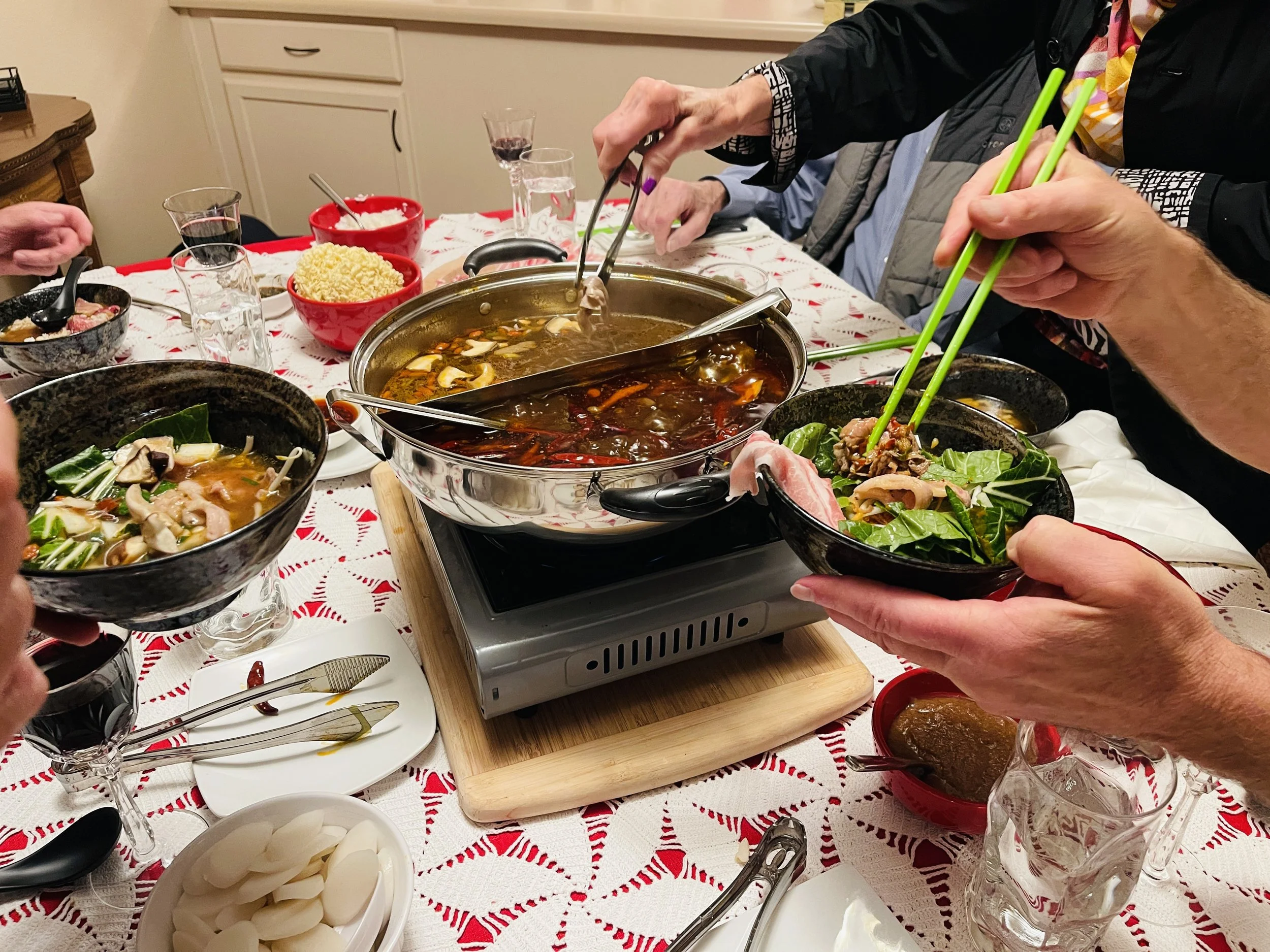 People sharing a hot pot meal, using chopsticks to pick ingredients from bowls and a large central pot on a portable stove, with side dishes and drinks on a table with a red and white tablecloth.