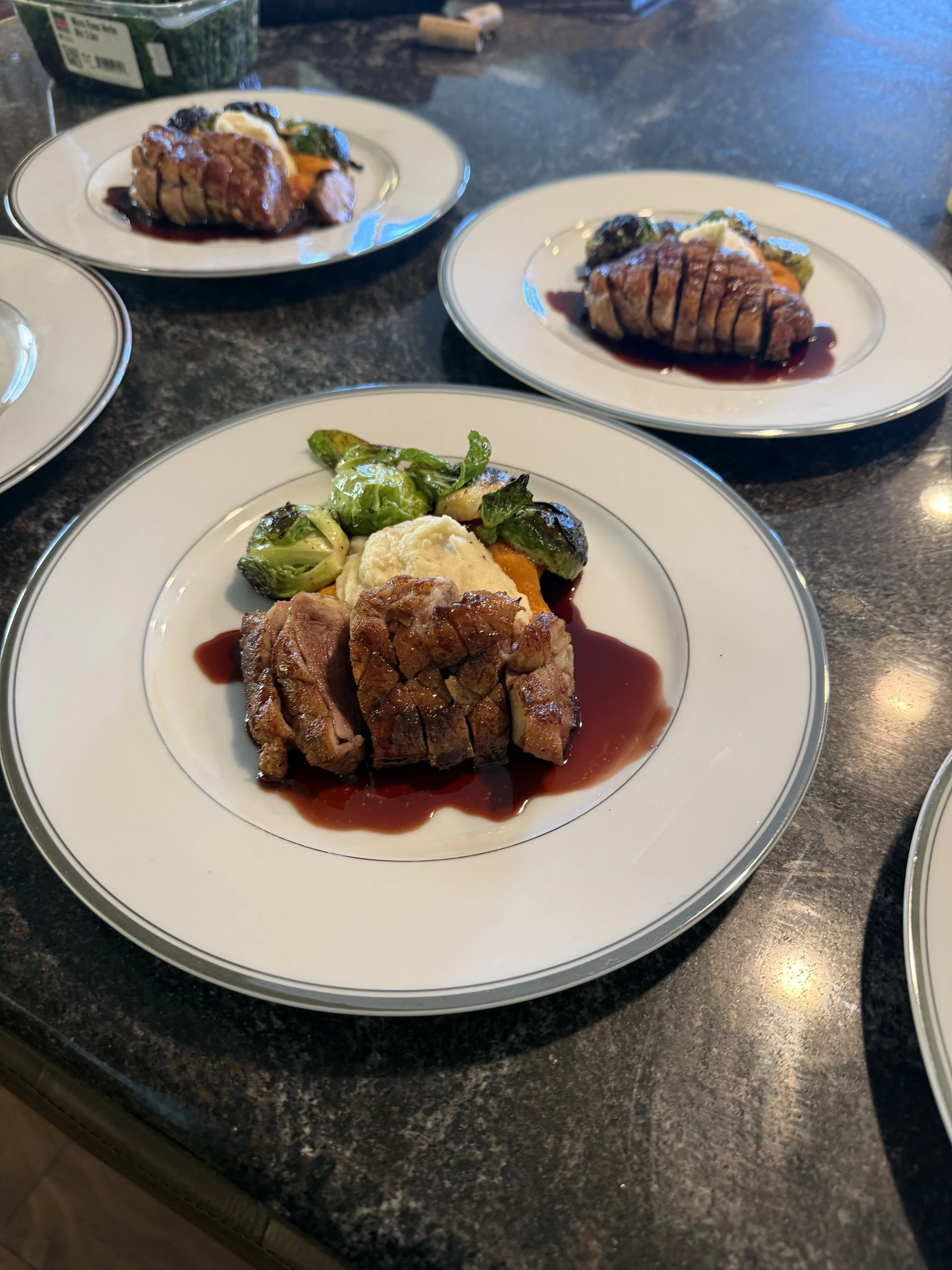 A plate with sliced roasted meat, Brussels sprouts, mashed potatoes, and a dark sauce garnish on a dark countertop.