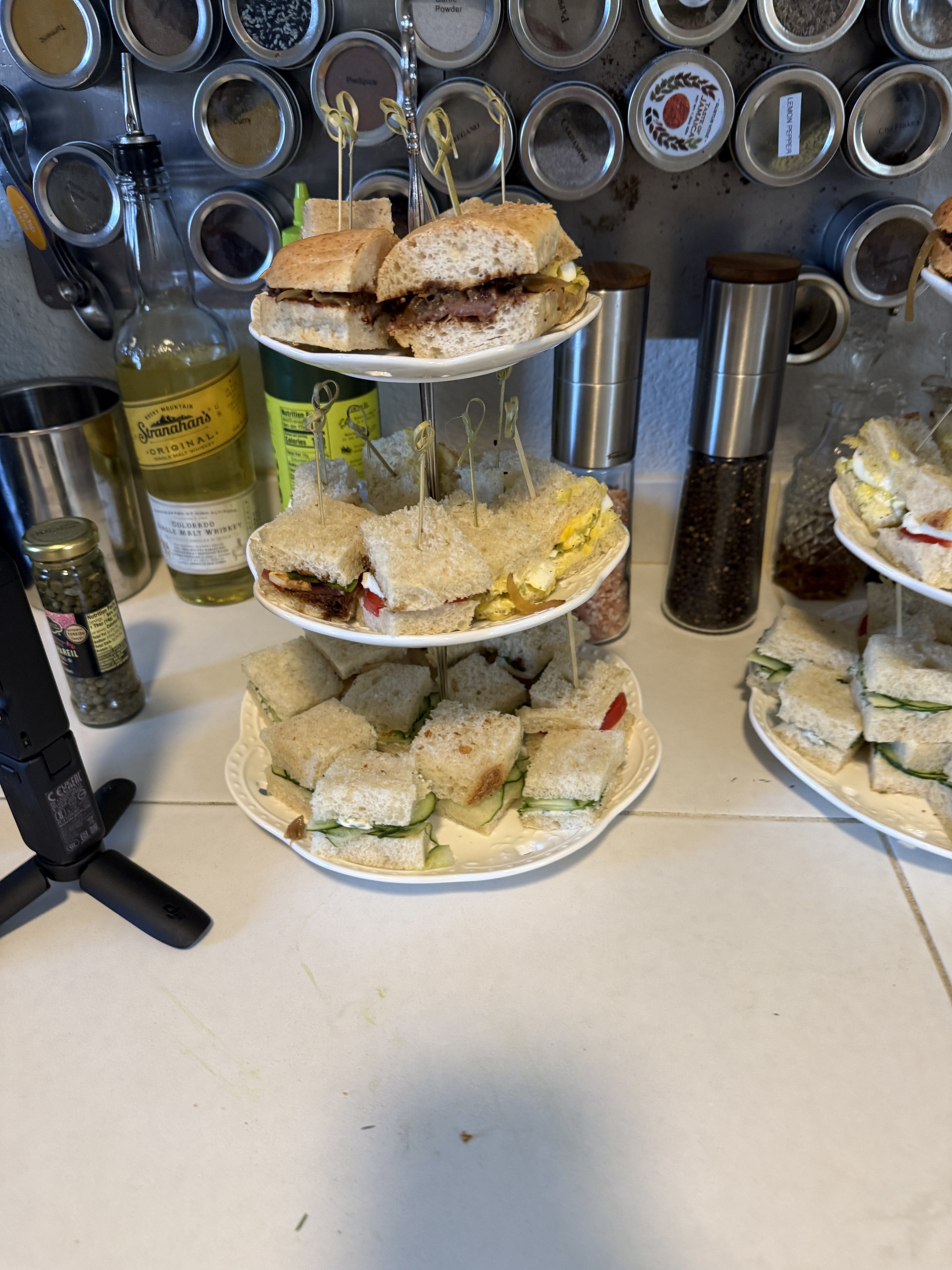 Three-tiered platter with assorted tea sandwiches and mini croissants, set on a counter with spice jars and condiments in the background.