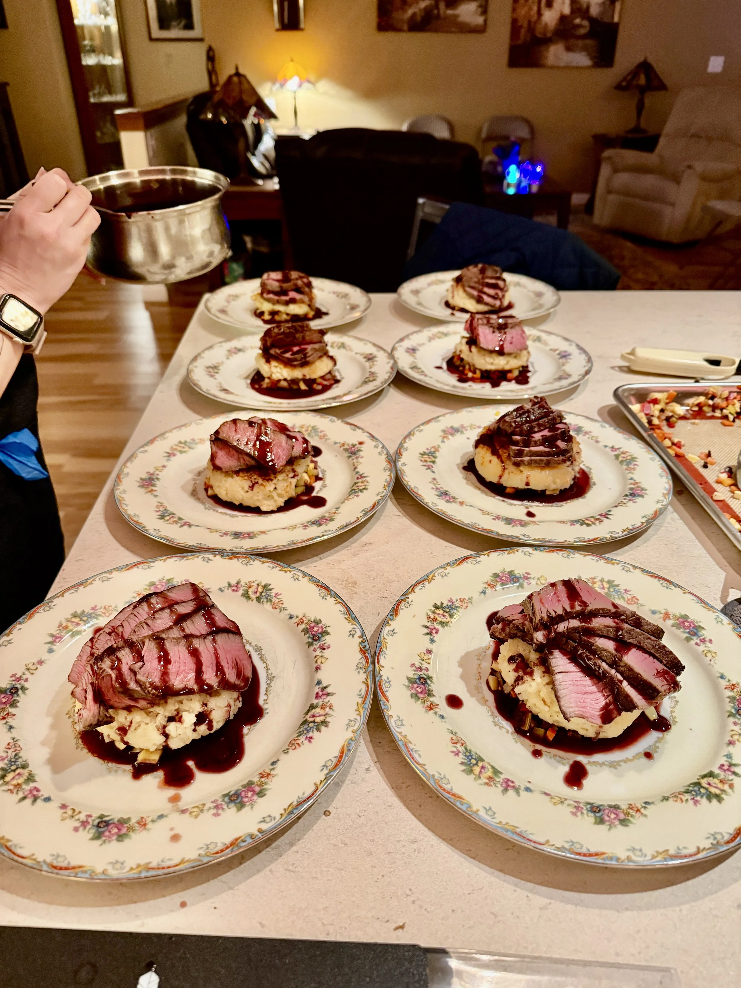 Several plates with slices of steak and mashed potatoes topped with gravy on a table, with a person pouring sauce from a saucepan.