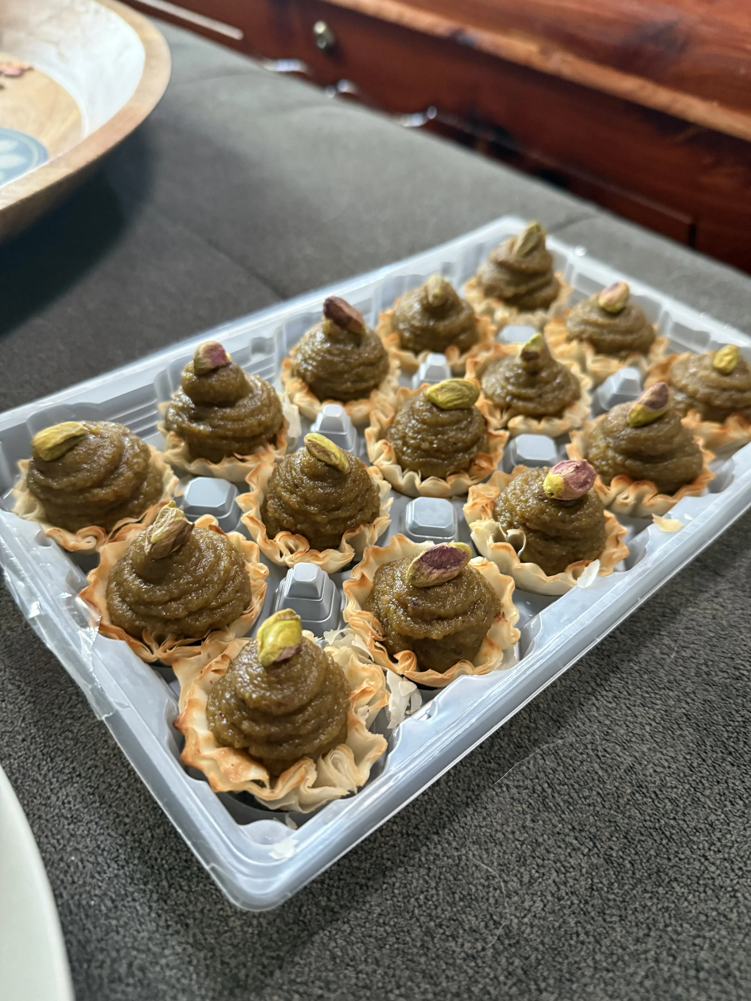 Small desserts with dollops of brown topping and pistachio nuts on top, arranged in flower-shaped paper cups on a plastic tray.