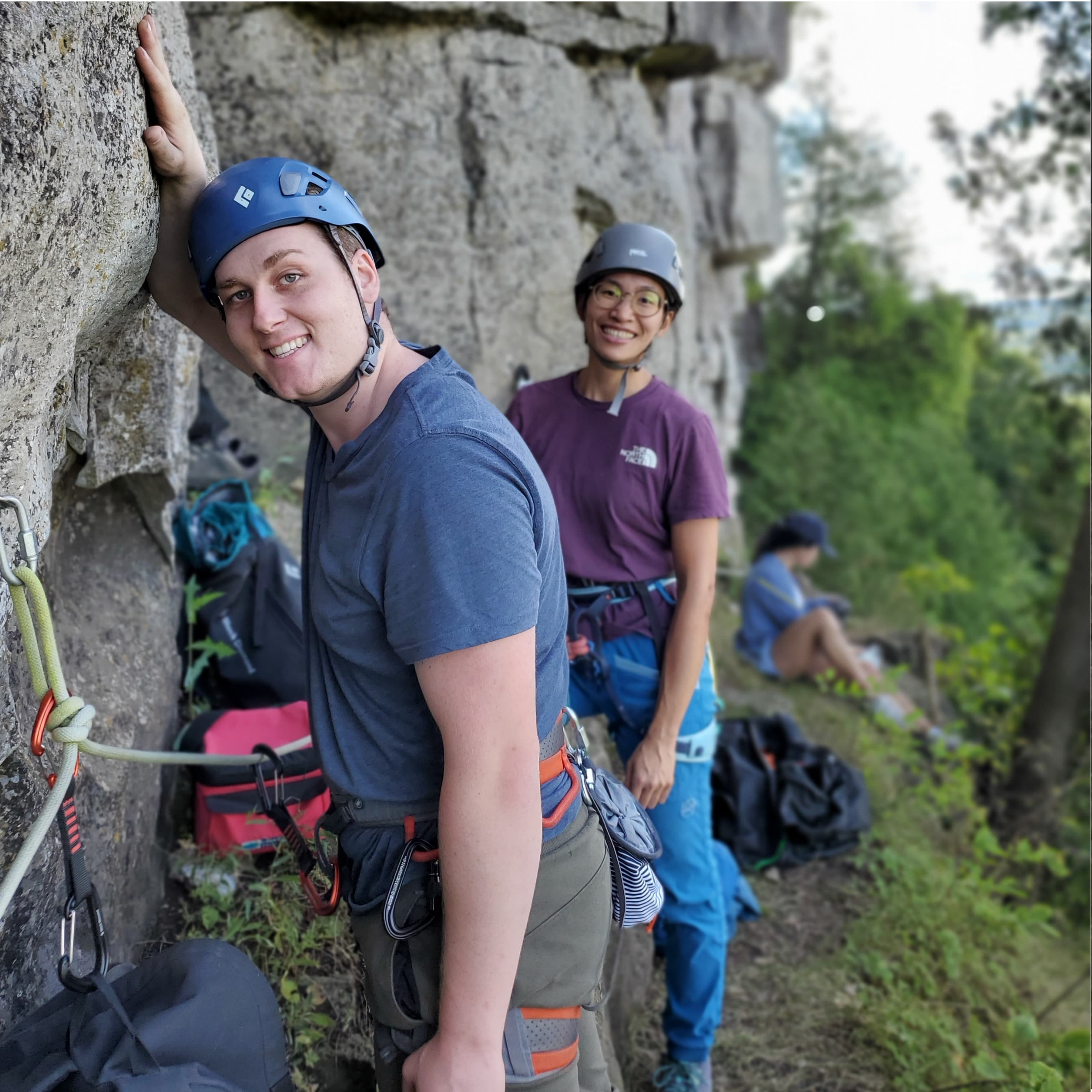 Two young climbers wearing helmets and climbing gear, standing on a rock face outdoors, smiling at the camera, with a person sitting in the background near trees and backpacks.