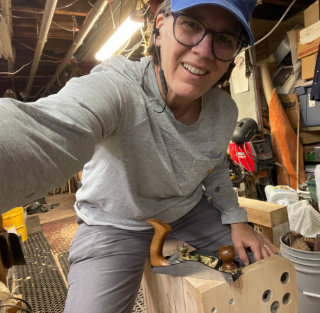 Person smiling in a basement workshop, wearing glasses and a blue cap, taking a selfie while working with a hand plane on a piece of wood.