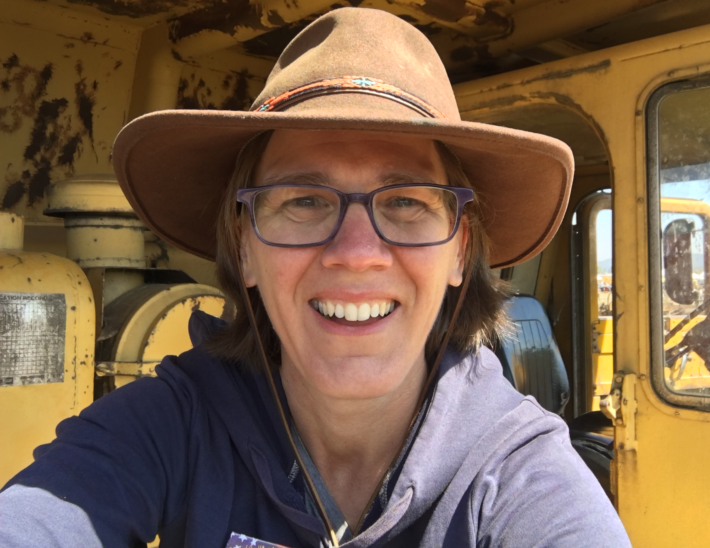Person smiling with glasses and wide-brimmed hat inside a yellow industrial vehicle or machinery