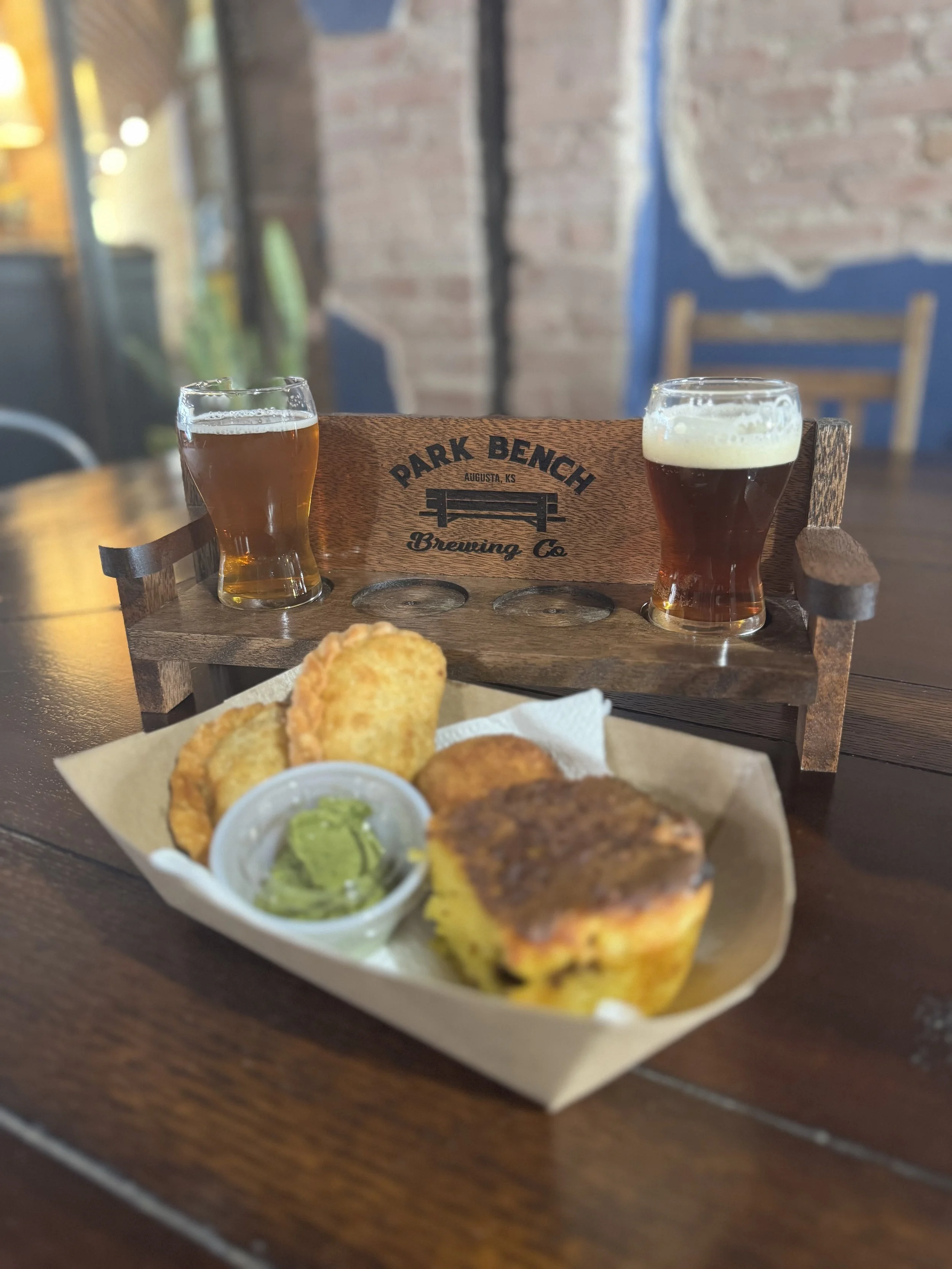Two glasses of beer on a wooden tray at Park Bench Brewing Co., with a plate of fried food and dipping sauce on a wooden table in a rustic setting.