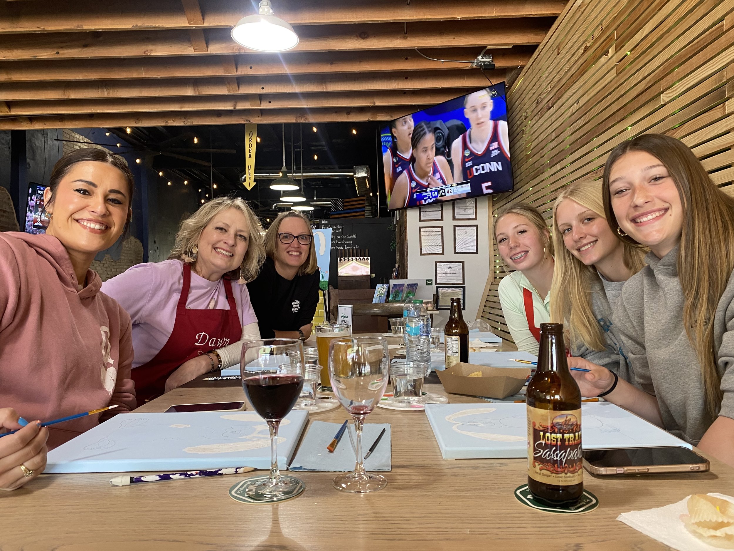Group of seven women smiling and sitting around a table with drinks and food in a casual restaurant, watching a basketball game on a TV mounted on the wall.