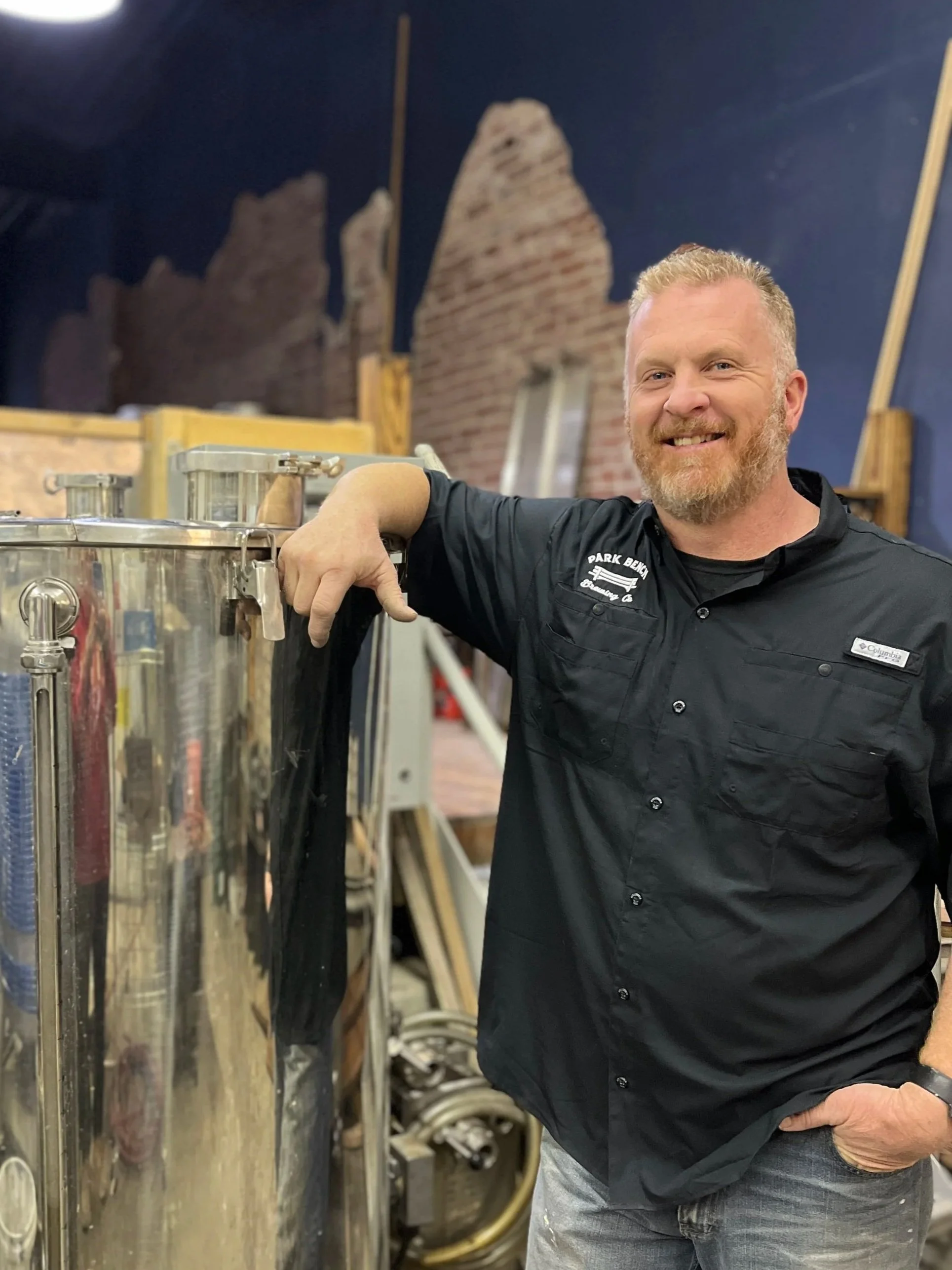 A man with a beard and short blonde hair smiling, wearing a black shirt with a logo, standing indoors beside a large piece of brewing equipment in a room with unfinished brick walls and wooden support beams.