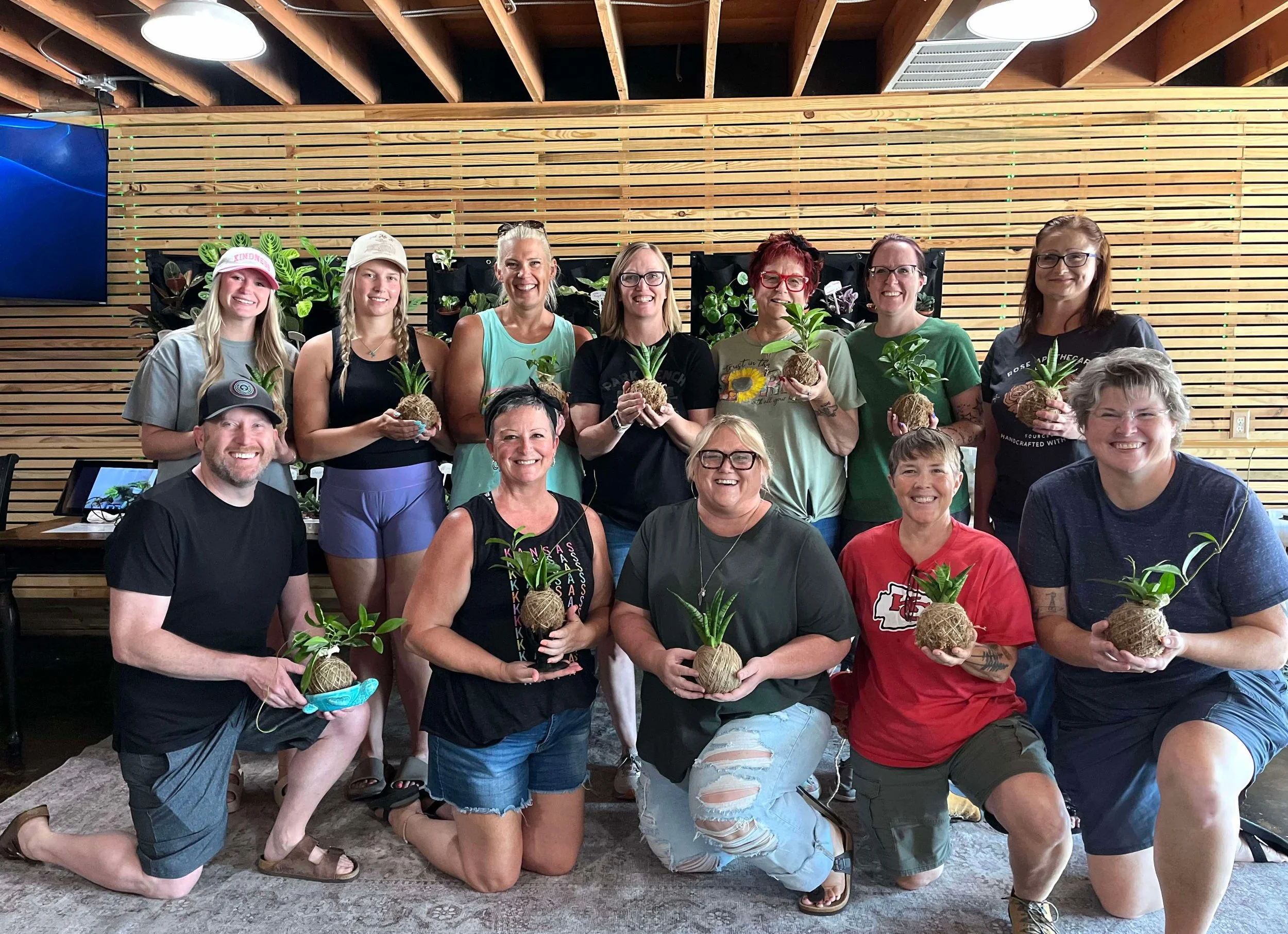 Group of eleven people holding plants in a room with wooden wall decor.