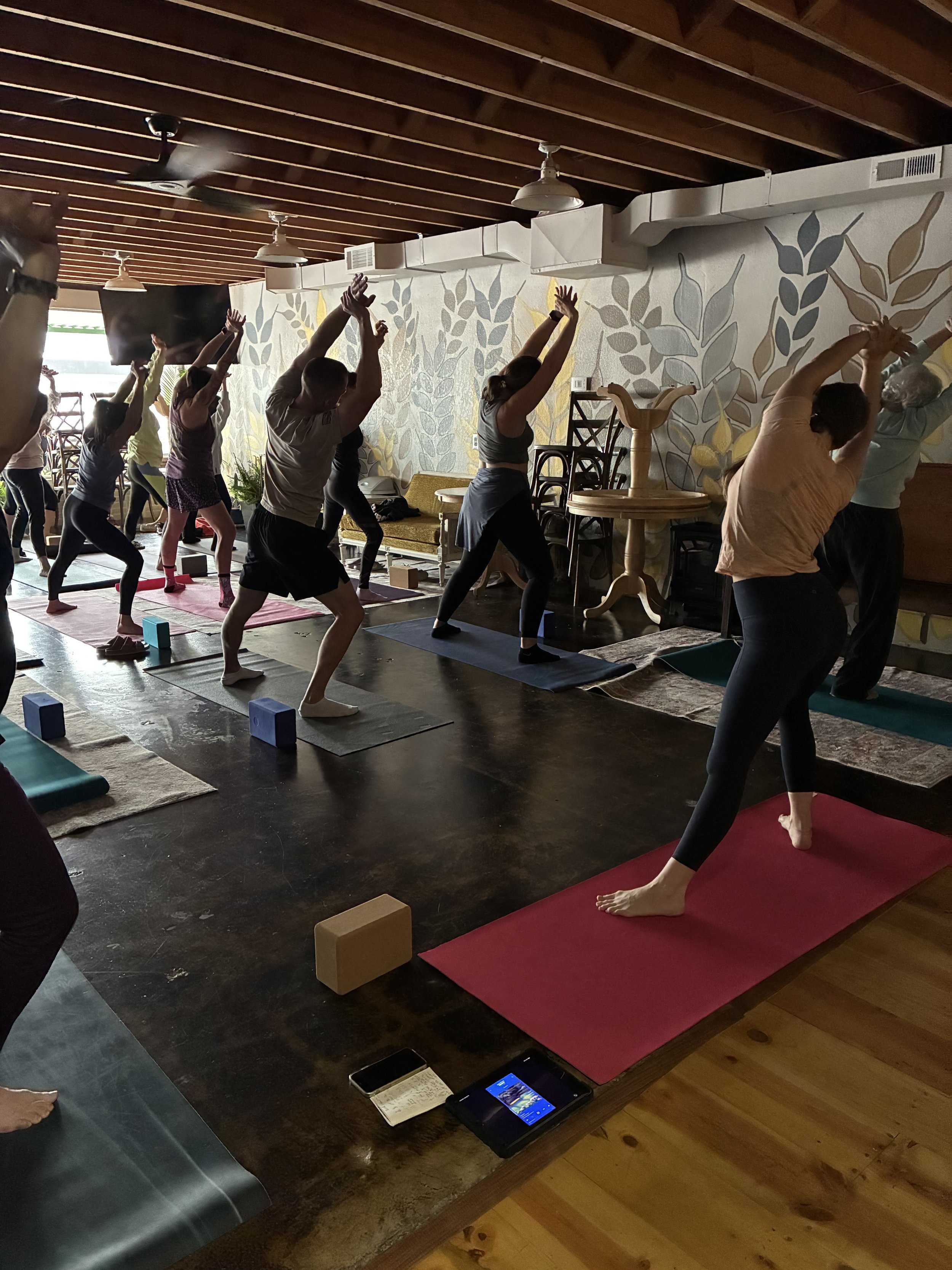 Group of people participating in a yoga class, performing side stretches on yoga mats inside a room with wooden ceiling beams and a decorative leafy wall.