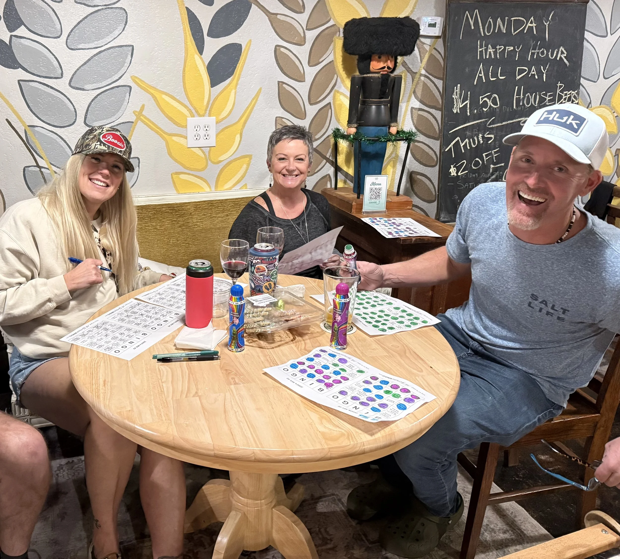 Three people sitting around a wooden table at a restaurant playing bingo, with drink cans and glasses, smiling and holding bingo cards, in front of a colorful leaf-patterned wall and a chalkboard with a special offer.