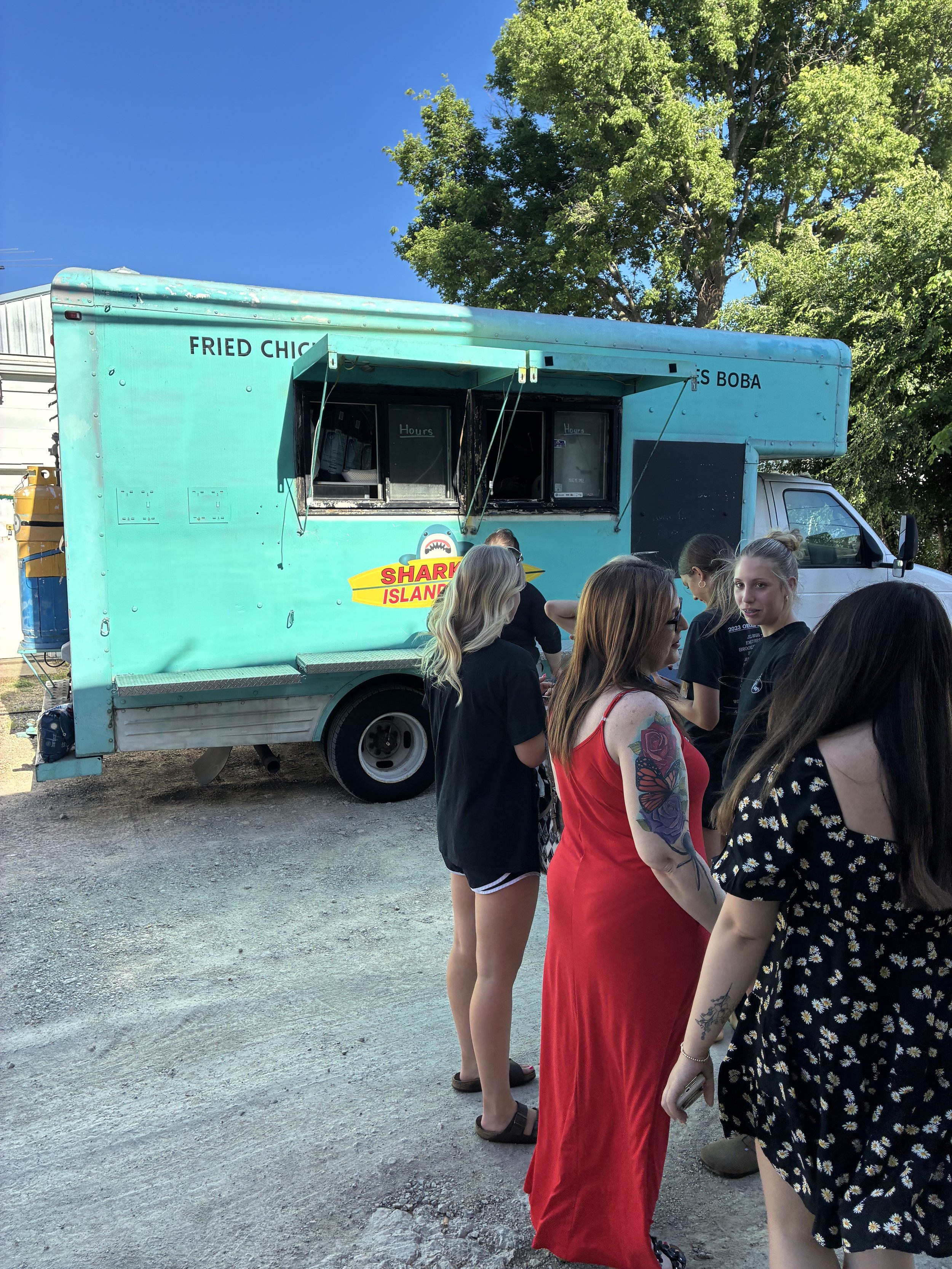 People waiting in line at a teal food truck with a sign indicating fried chicken and sushi boba, under a large green tree on a sunny day.