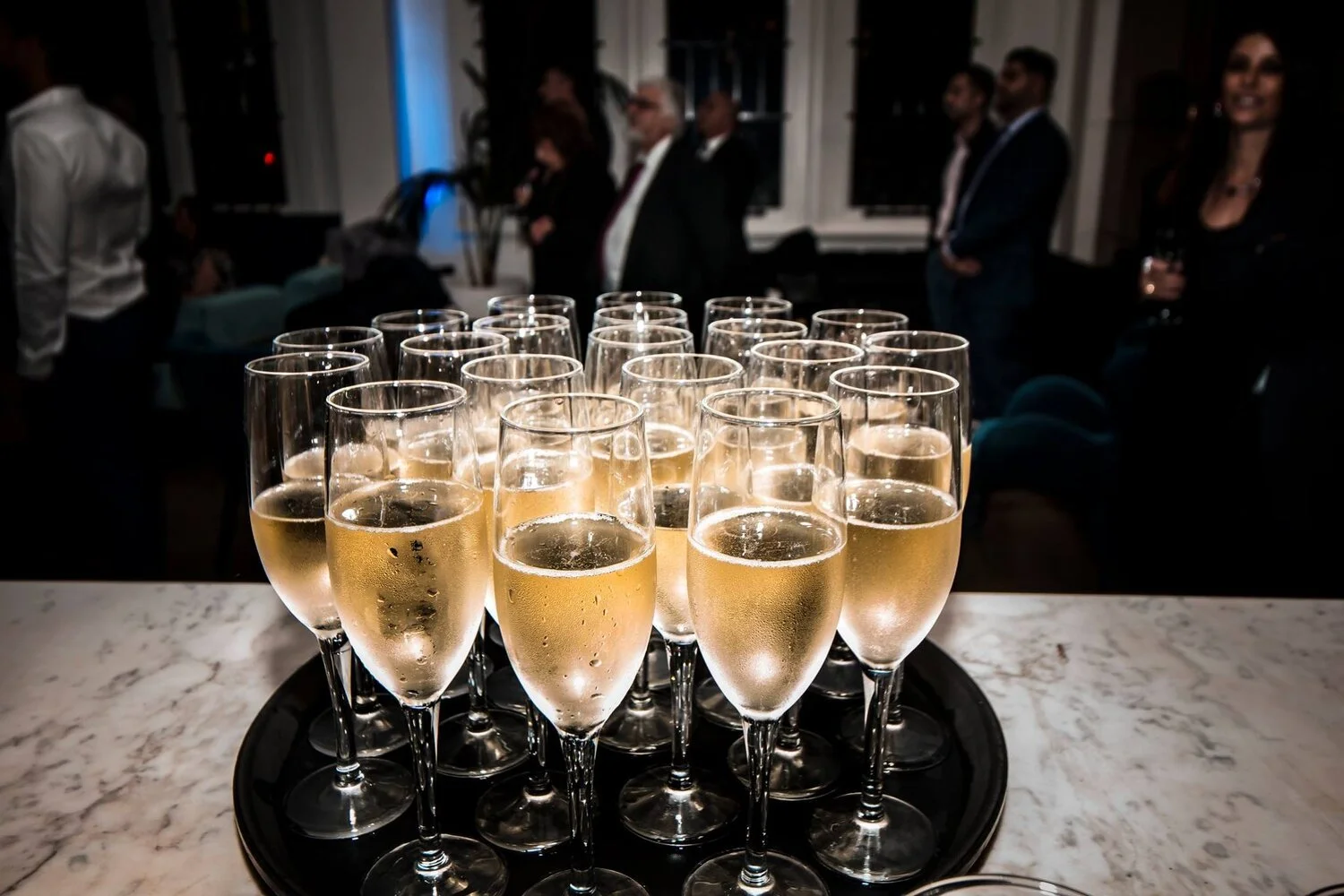 Tray of champagne glasses filled with bubbly on a marble table in a dimly lit reception room, with guests mingling in the background.
