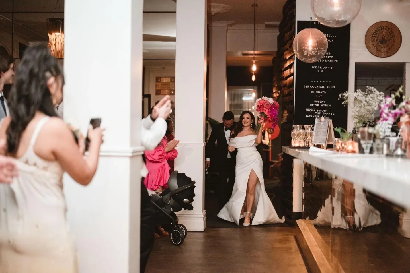 Woman in wedding dress holding colorful bouquet walking into reception area while guests watch and take pictures.