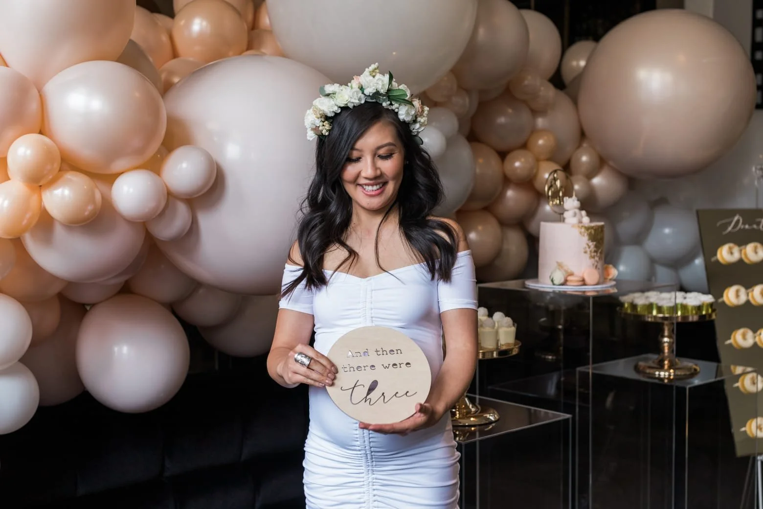A woman in a white dress and floral headband holding a sign that reads "And then there were three" at a celebration with balloons and cake.