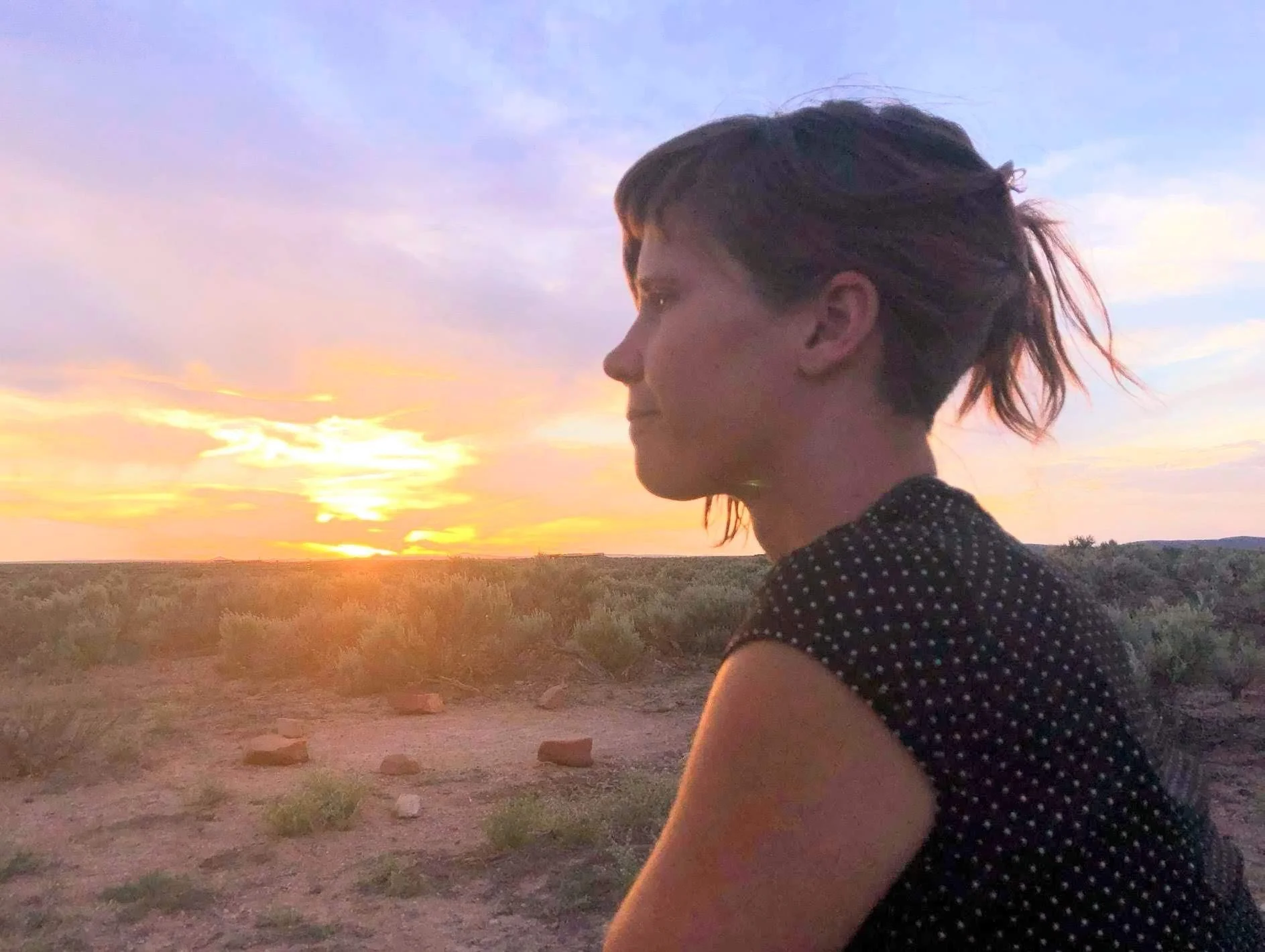 A woman with short brown hair in a ponytail sitting outdoors during sunset, wearing a black top with white polka dots, with open landscape and bushes in the background.
