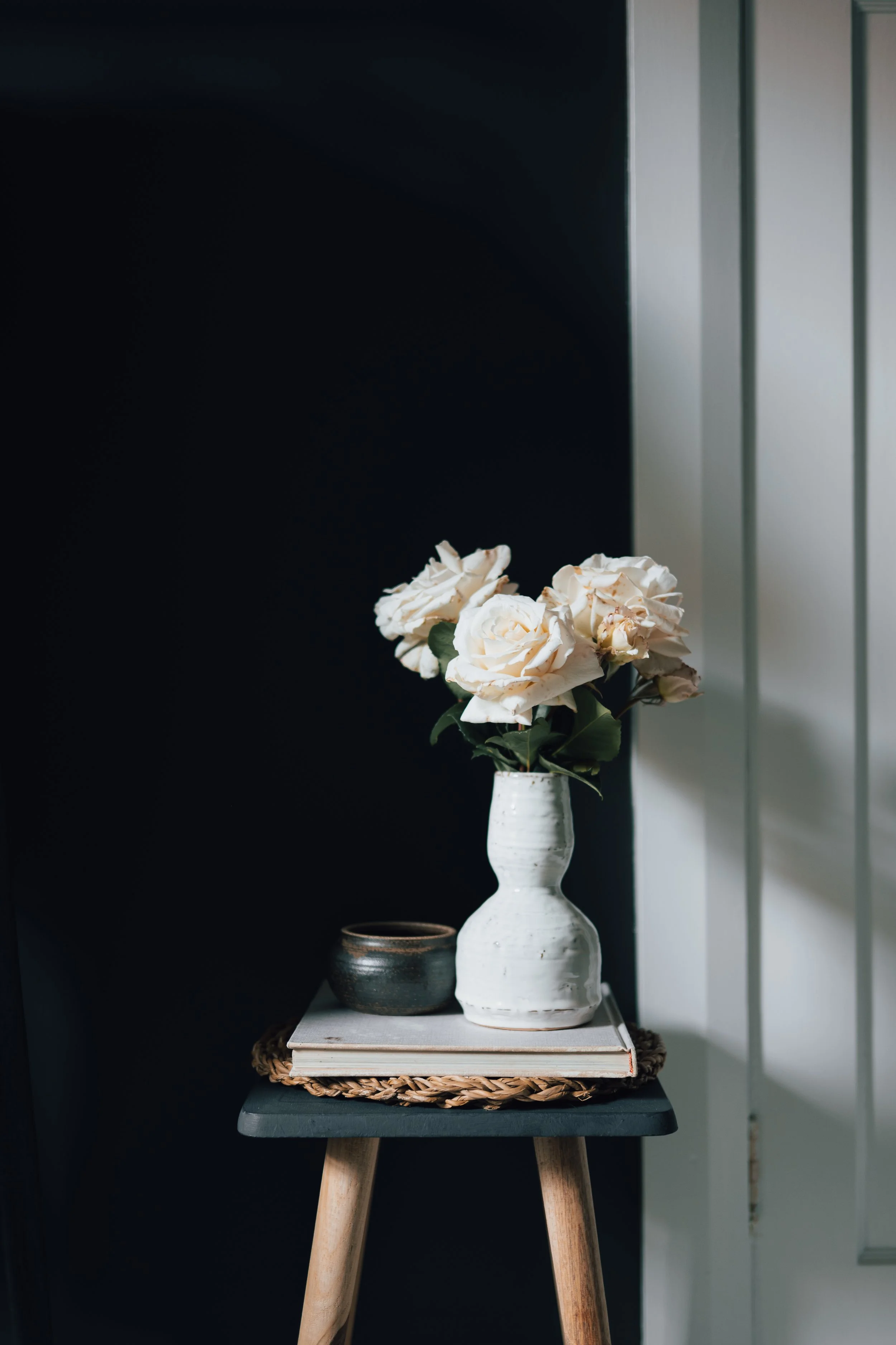 A white ceramic vase with white and light pink roses, a smaller dark ceramic bowl, and a book on a woven placemat on a wooden stool against a dark wall and light-colored paneling.